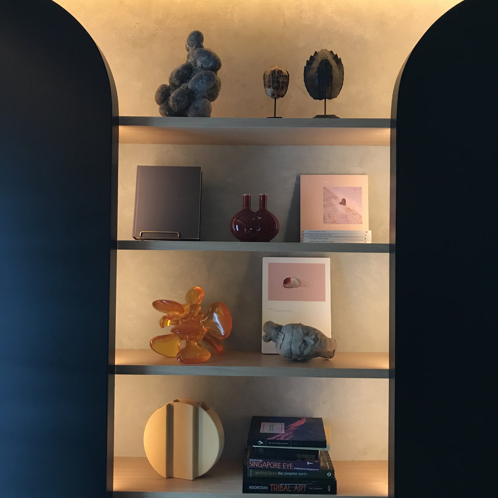 A lit alcove of shelves holding ceramics, books and small objects in amber and blue