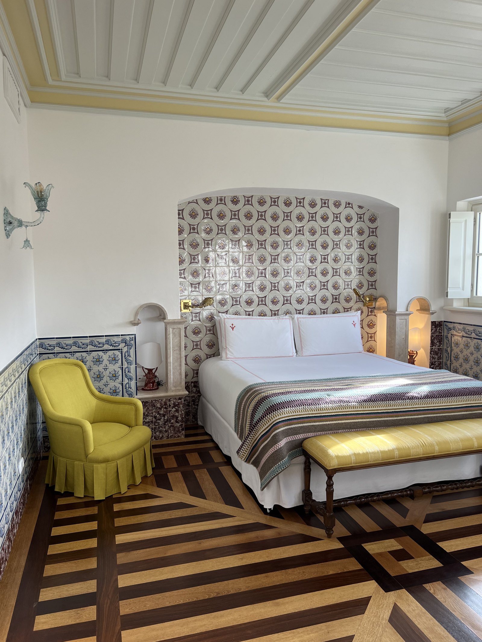 A bedroom at Vermelho with a tiled headboard, a yellow armchair and a chevron parquet floor