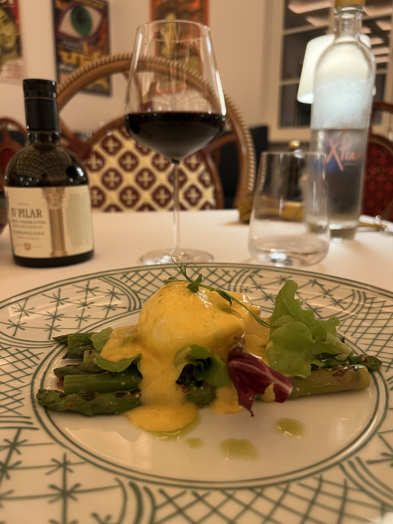 Eggs with hollandaise over asparagus on a green patterned plate, with a glass of red wine behind