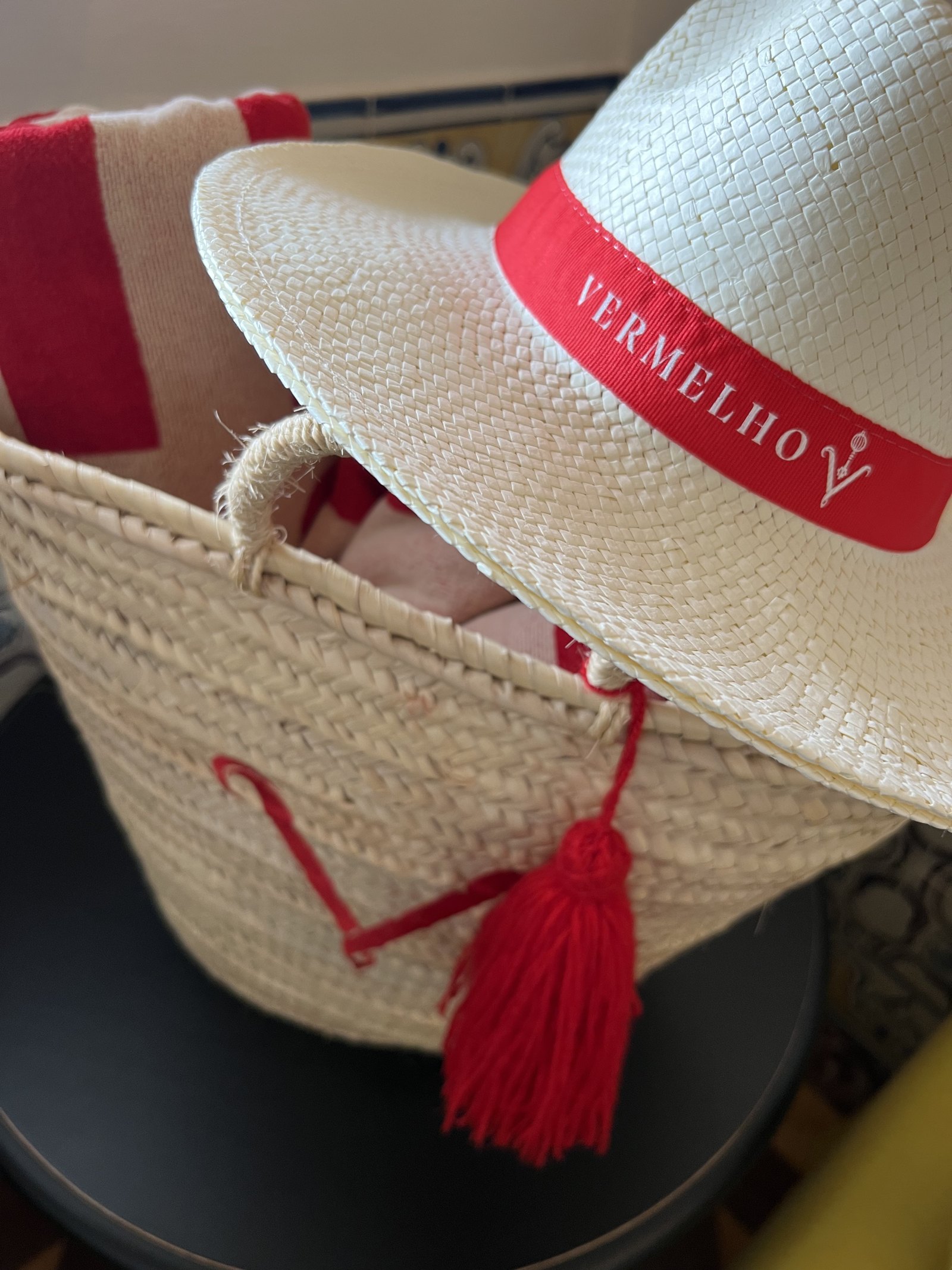 A straw basket holding a straw hat with a Vermelho ribbon and a red tassel
