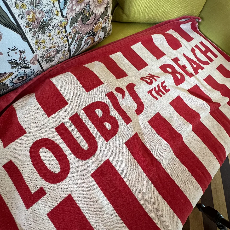 A red and white striped beach towel printed Loubi's on the Beach, folded on a green cushion