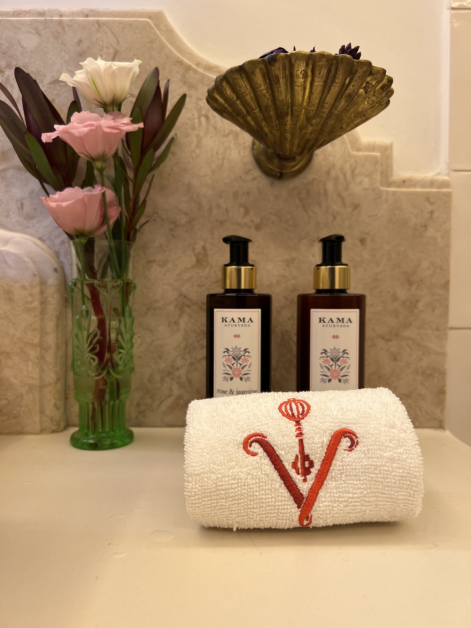 Kama Ayurveda bottles and a rolled towel embroidered with a red V, beside flowers under a brass shell sconce