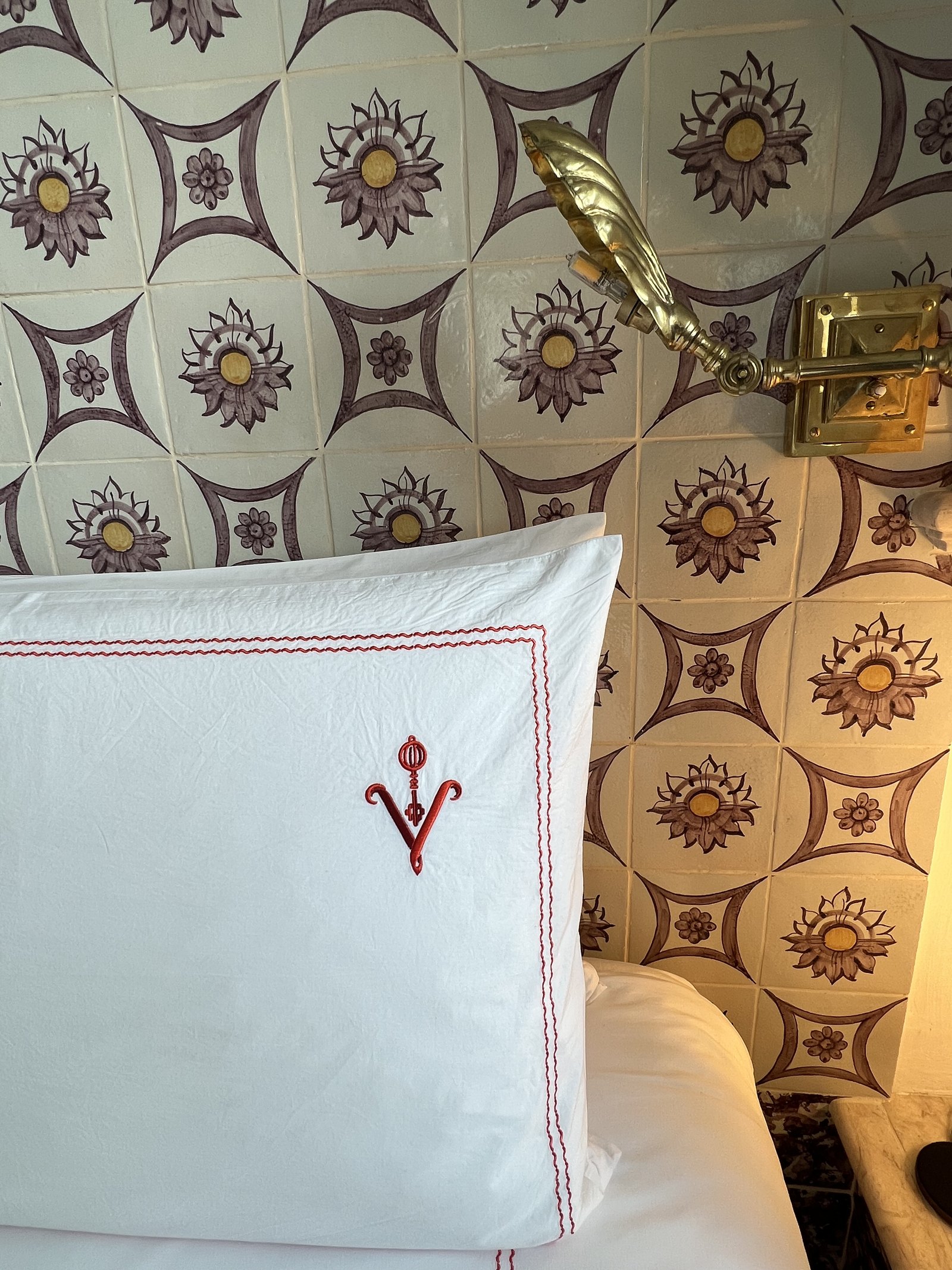 A white pillow embroidered with a red V against a wall of sun patterned azulejos, lit by a brass shell sconce