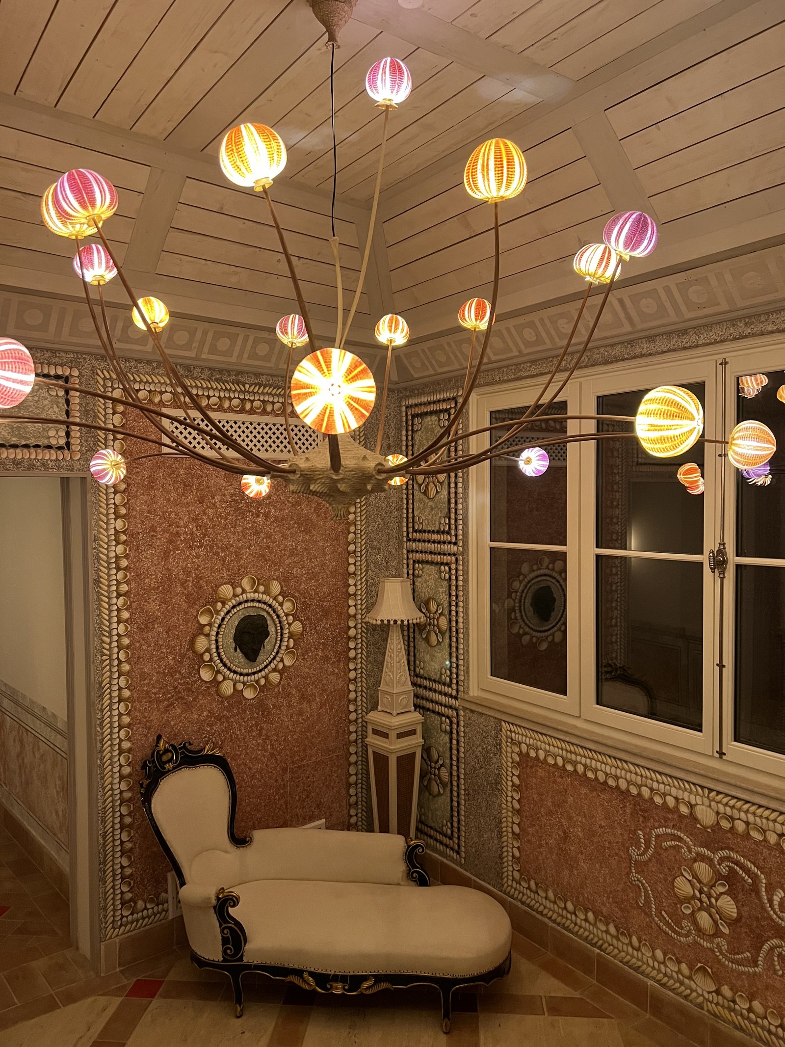 A salon with coloured glass pendant lanterns, a chaise longue and frescoed pink walls