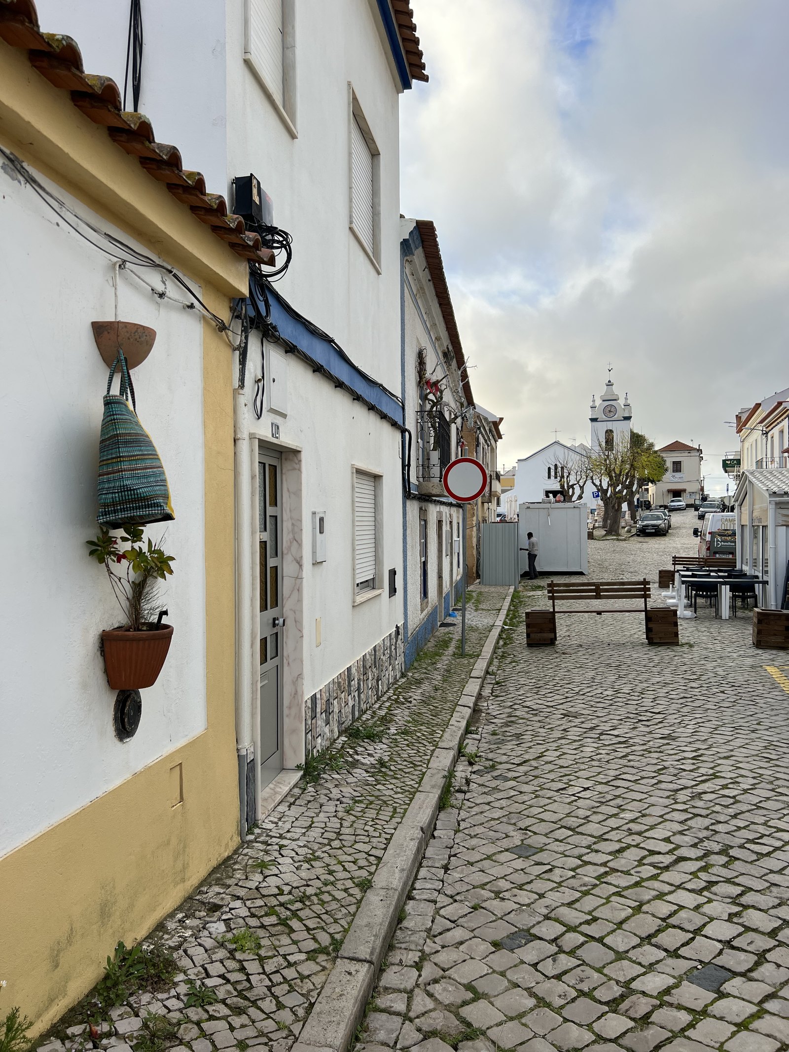 A cobbled street of whitewashed houses in Melides with a hanging ceramic pot and the church tower beyond