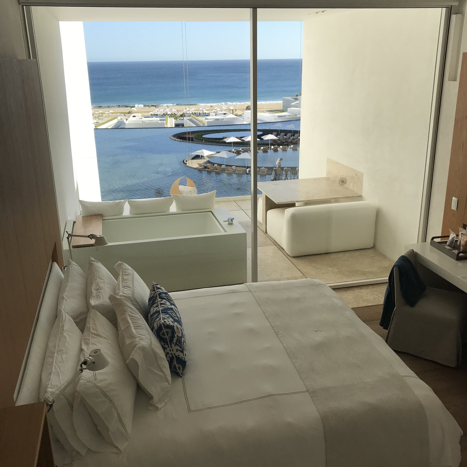 A bed with white linen and a blue patterned cushion at Viceroy Los Cabos, with a tub set against the window and the sea and pools beyond