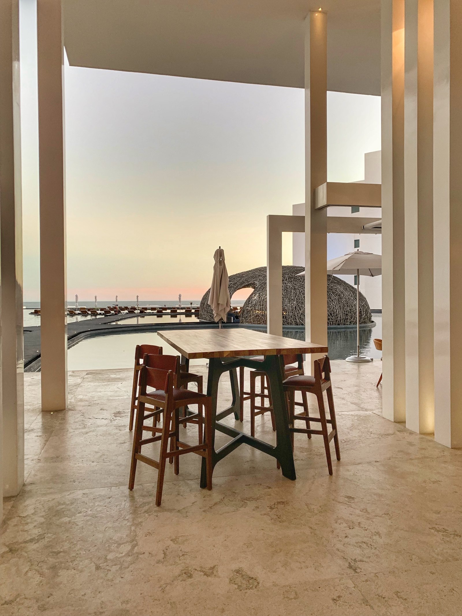 A view between white columns at Viceroy Los Cabos at sunset, with a bar table and stools in the foreground and the sea and the woven dome beyond