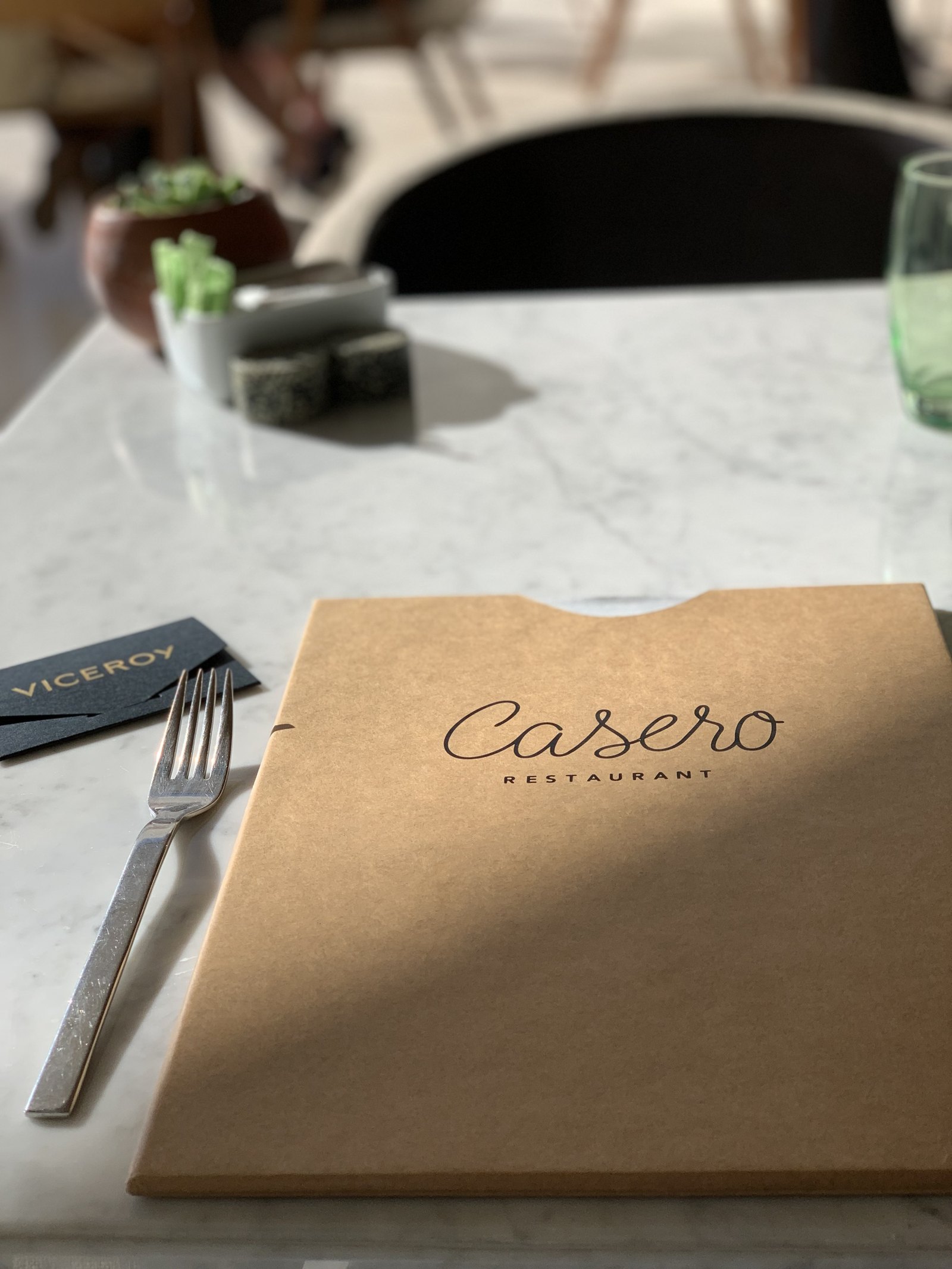 A menu in a brown card cover reading Casero Restaurant, lying on a marble table at Viceroy Los Cabos beside a fork and a glass