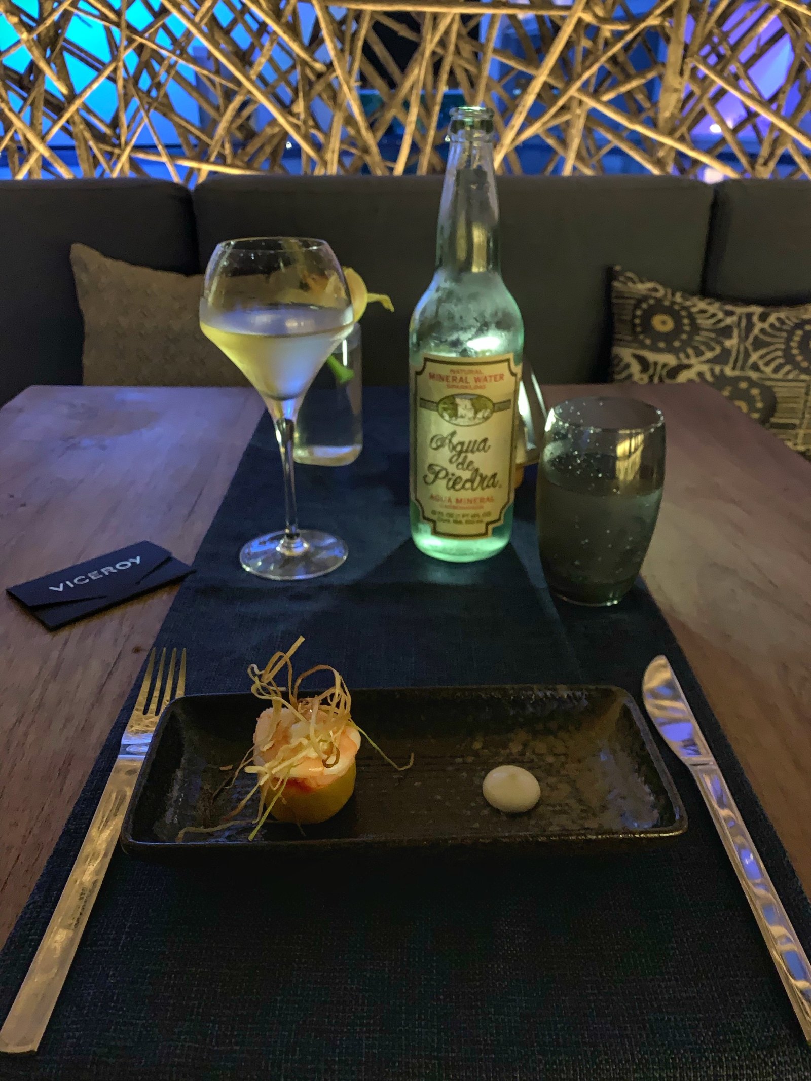 A small composed dish on a black tray at Viceroy Los Cabos with a glass of white wine and a bottle of mineral water on a dark table, the lit weave of the dome overhead