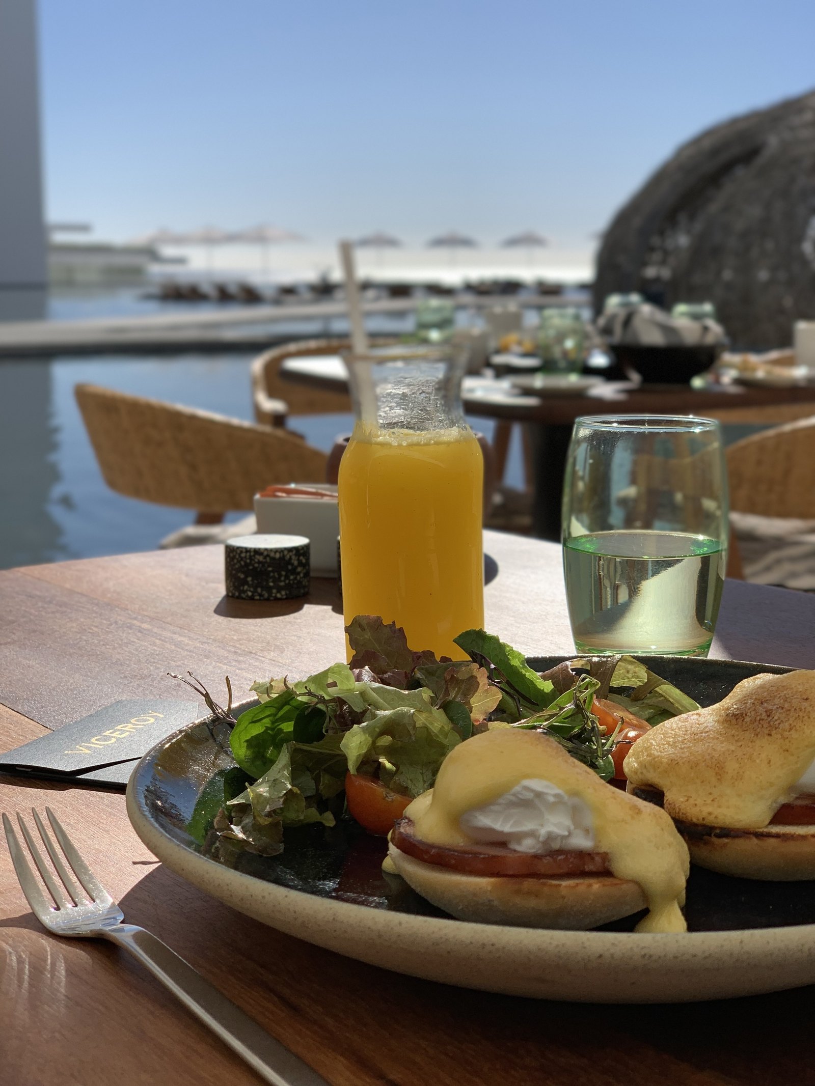 A plate of eggs on toasted bread with hollandaise and a green salad at Viceroy Los Cabos, with a bottle of orange juice and a glass of water behind it on a timber table