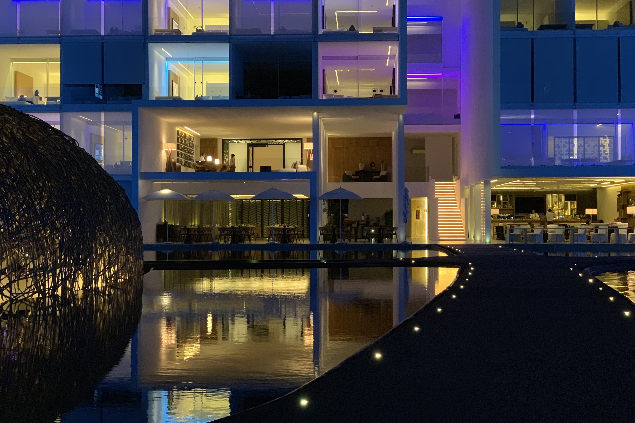 The facade of Viceroy Los Cabos lit violet and magenta after dark, doubled in the still black water in front of it, with the woven dome dark in the foreground