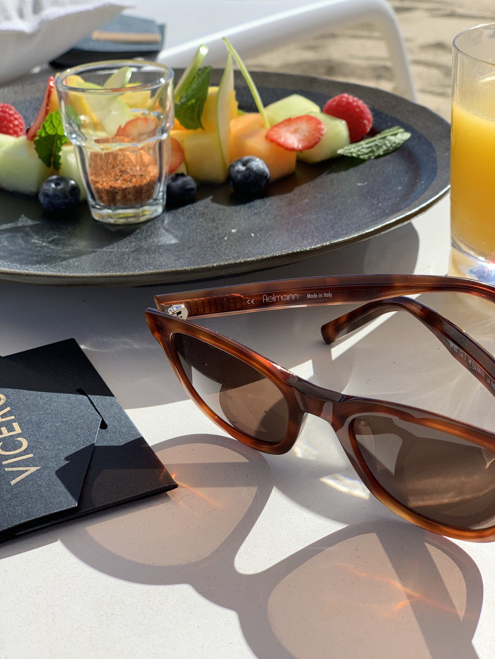 A plate of cut melon, papaya and berries with a small glass of granola at Viceroy Los Cabos, beside a pair of tortoiseshell sunglasses on a dark folder
