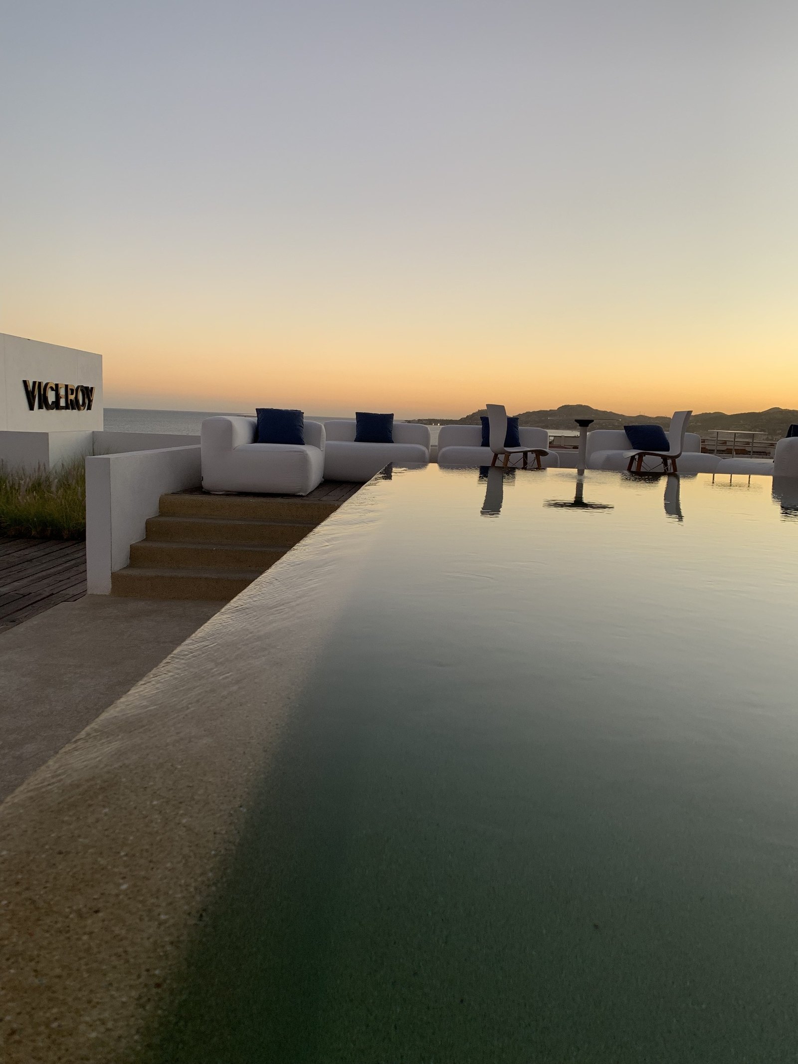 The long infinity pool at Viceroy Los Cabos at sunset, perfectly still, with the hotel lettering on a white wall at the left and the mountains across the bay