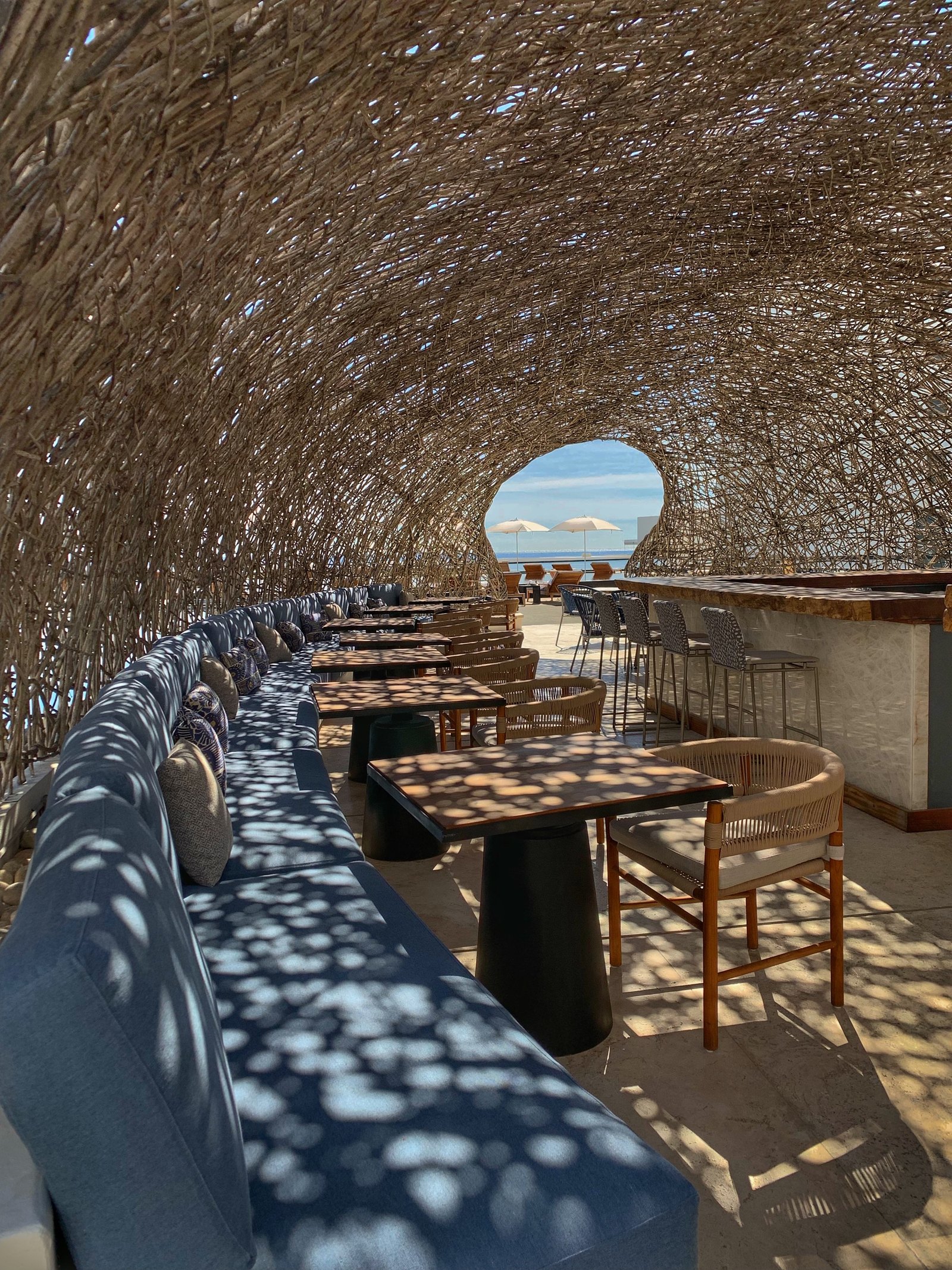 The interior of the woven dome at Viceroy Los Cabos with a long upholstered banquette and tables running down it, the sun through the branches casting a net of shadow across everything