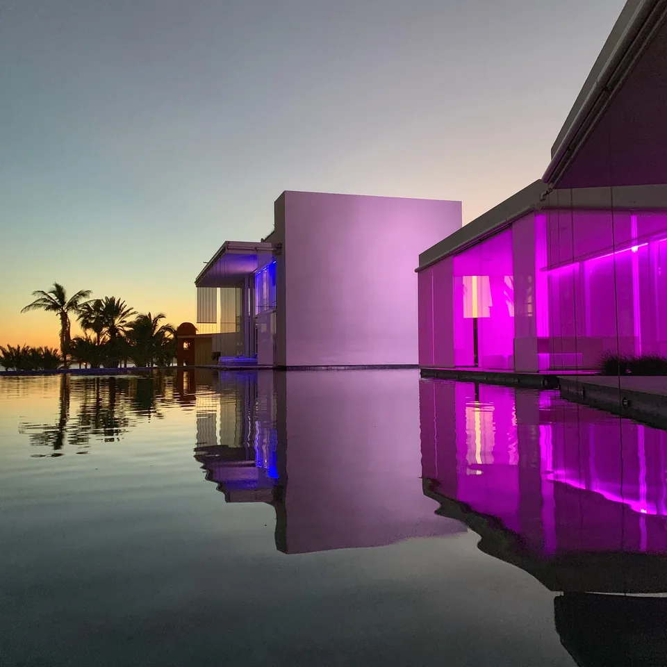 A wall of the hotel lit magenta after sunset at Viceroy Los Cabos, doubled in the still black water in front of it, with palms against the last light
