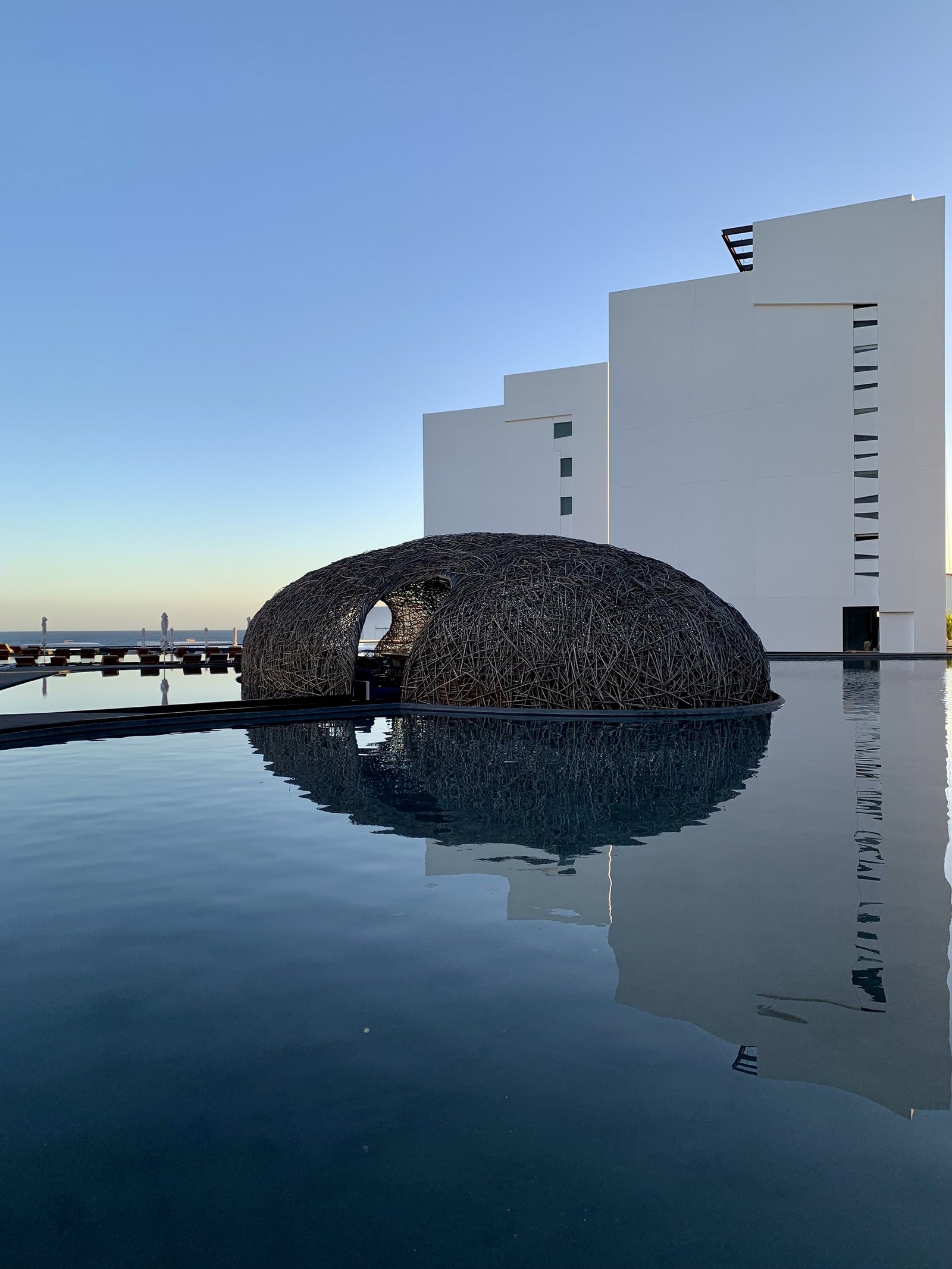 The woven nest of the restaurant at Viceroy Los Cabos sitting low in a sheet of black water, with the white stepped blocks of the hotel rising behind it