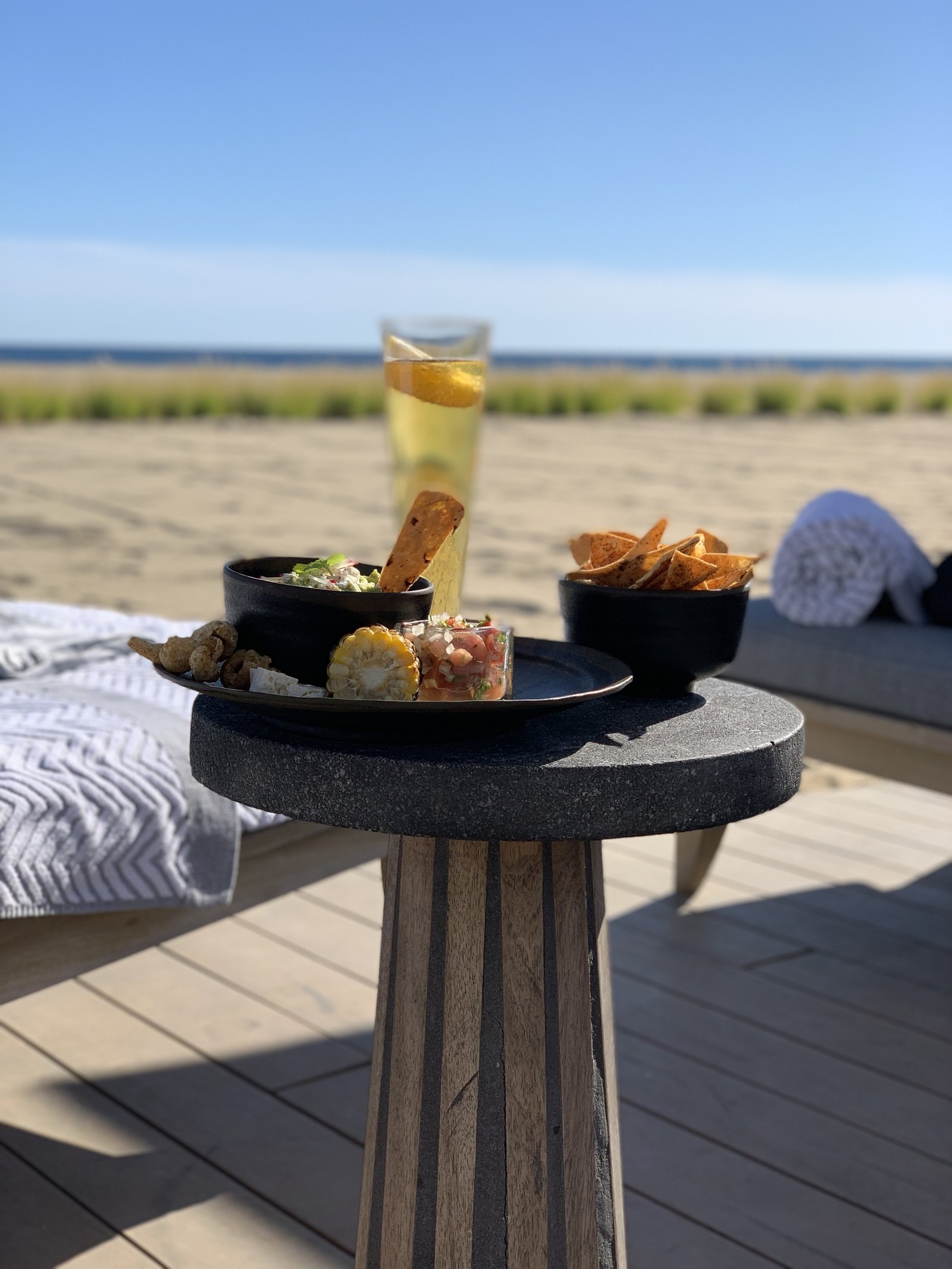 A dark stone table at Viceroy Los Cabos holding two small bowls of food and a tall glass of white wine, with a daybed, the sand and the sea behind