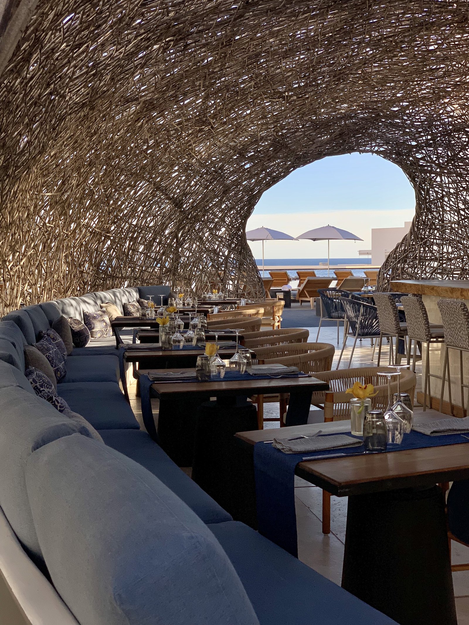 Set tables and blue banquettes inside the woven dome at Viceroy Los Cabos, with a round opening at the end framing the sea and a parasol