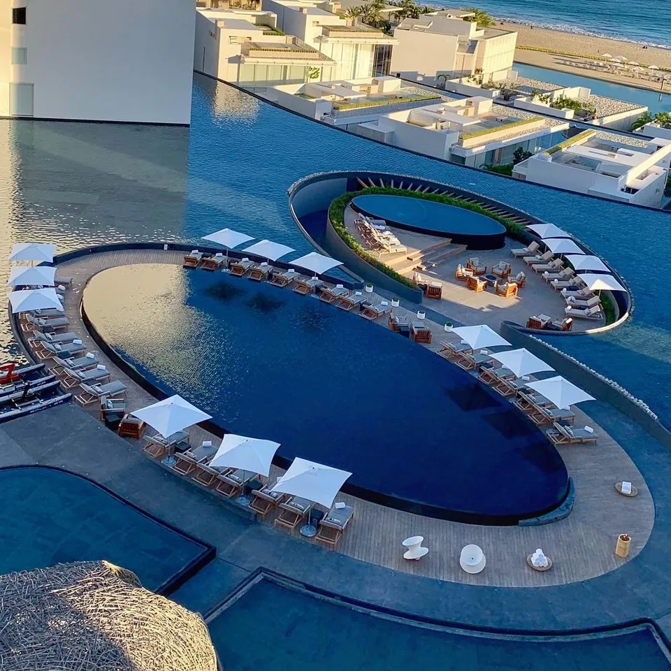 The oval pool at Viceroy Los Cabos seen from above, ringed by white loungers and parasols, with the sea and the white blocks of the hotel behind