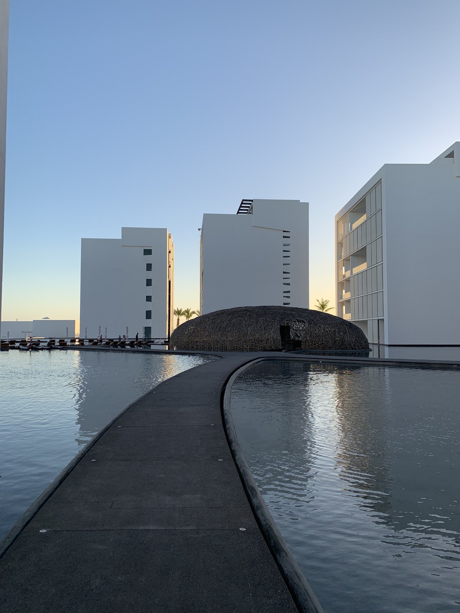 The dark water at Viceroy Los Cabos curving away in a long arc with the woven dome of the restaurant at the end of it and the white blocks of the hotel behind