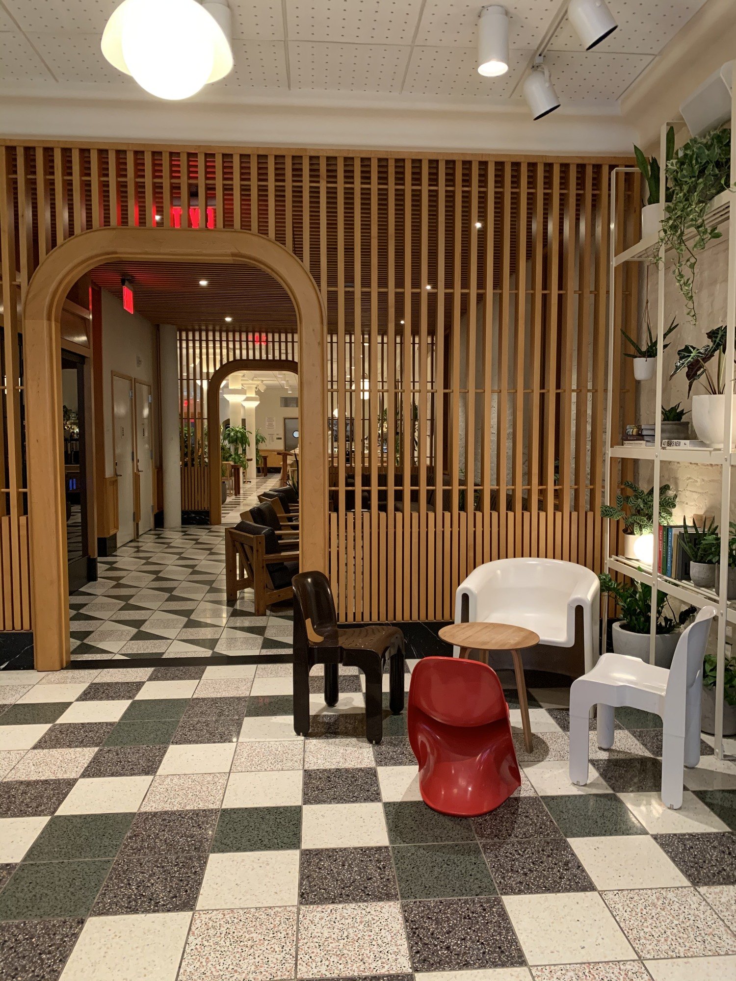 An oak-slatted screen with a tall arch cut through it, a red moulded stool and a white chair on a green and white checkerboard terrazzo floor