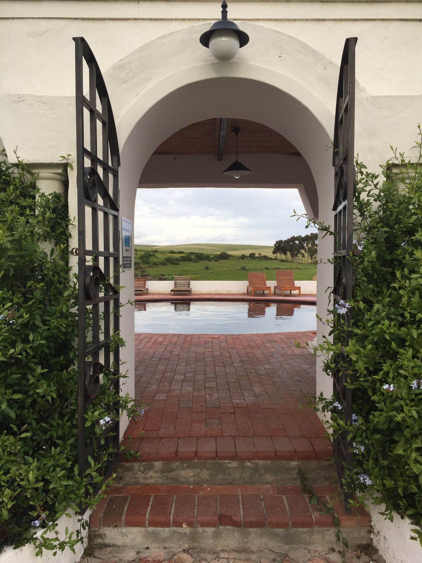 An open wrought-iron gate under a white arch, with a brick terrace, a round pool and timber loungers beyond, and green hills behind