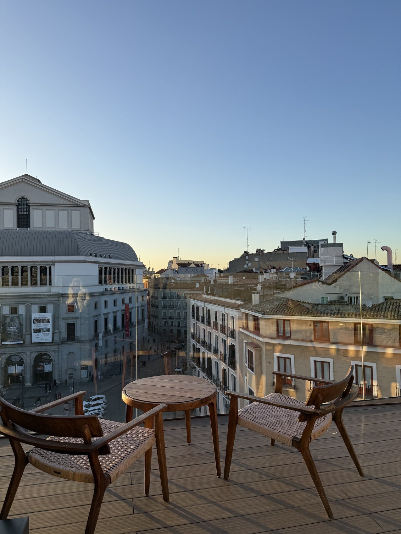 A balcony with woven chairs and a small table looking over the opera house and the rooftops at dusk
