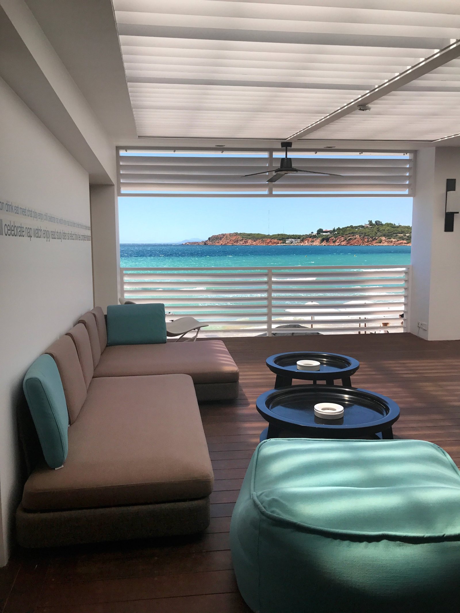 A beach club lounge with a long banquette, a turquoise floor cushion and two round black tables on a dark timber deck, open through white louvres to a turquoise sea and a red headland, a frieze of small words running along the wall