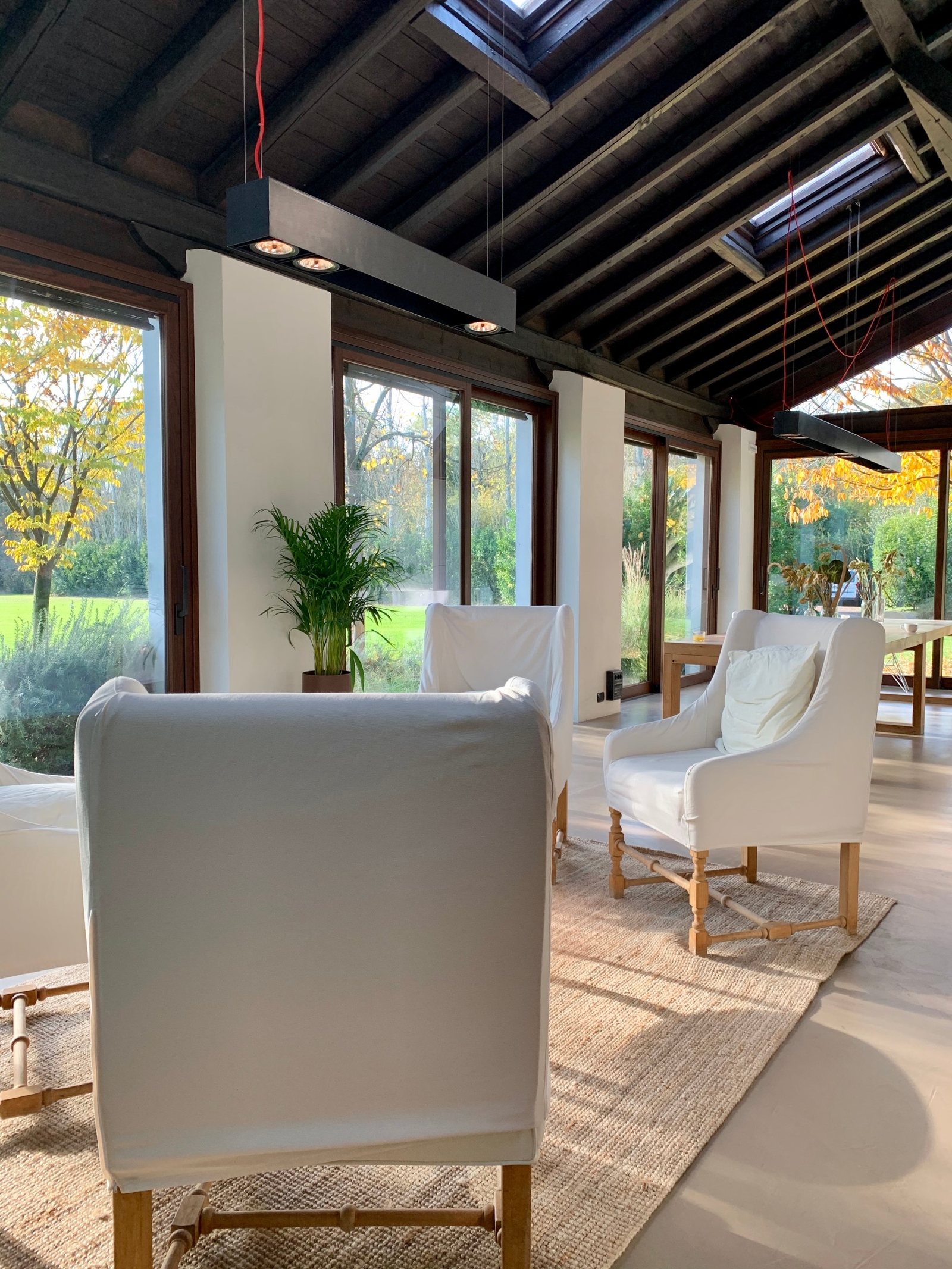 A converted barn sitting room with white slipcovered armchairs on a jute rug under a dark beamed roof, full-height windows down both walls onto a lawn and yellowing trees