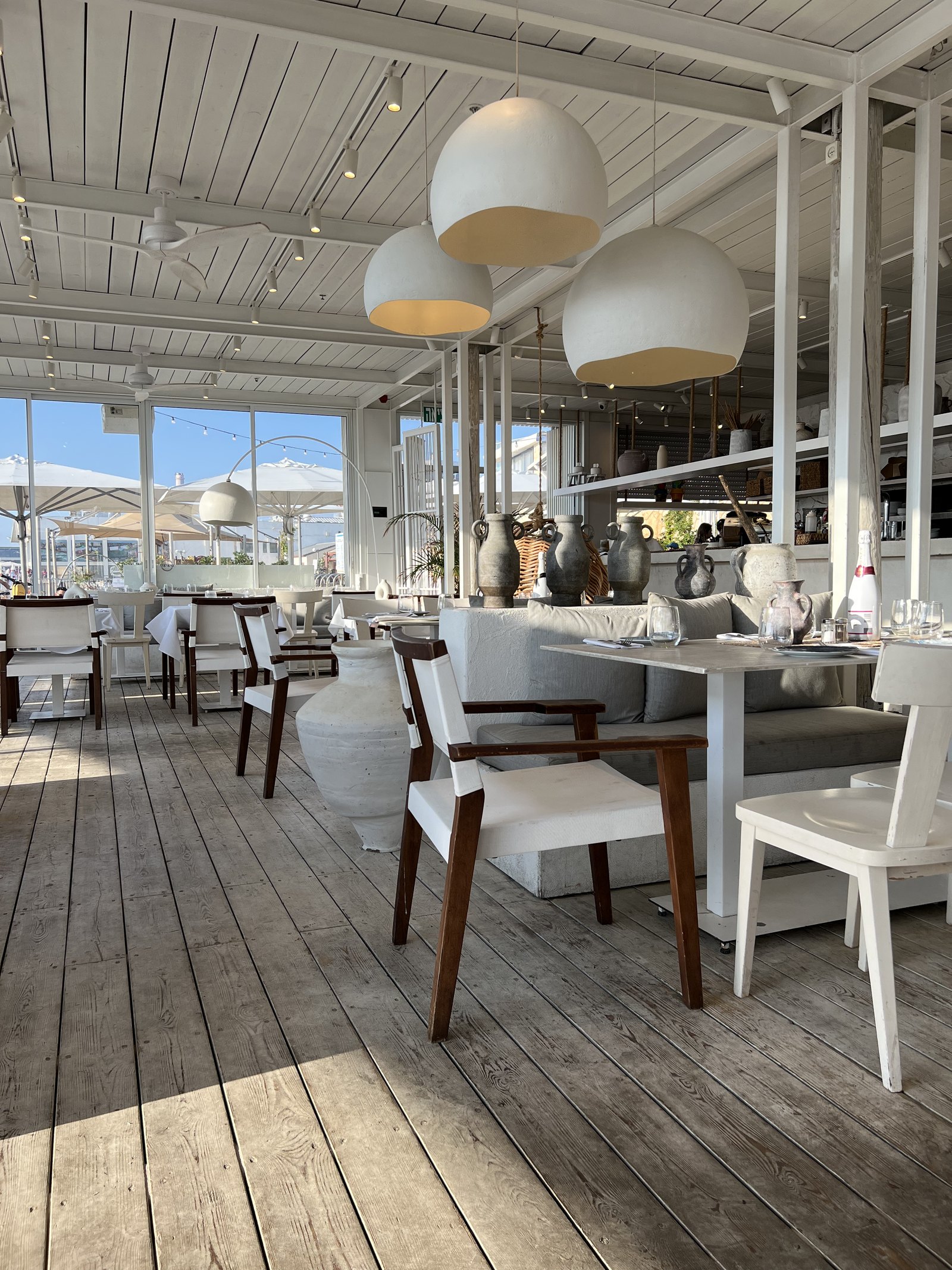 A beach club dining room under white globe pendants, with timber floors and pale chairs open to the sand