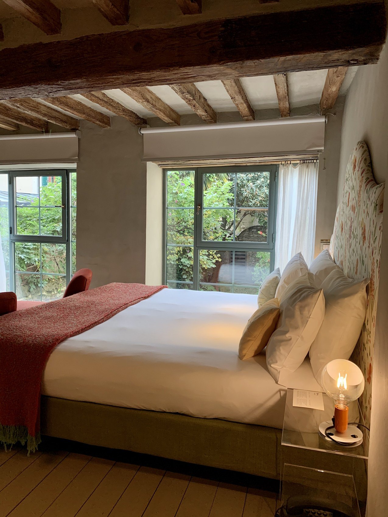 A bed piled with cushions under a rough beamed ceiling, tall windows onto a green garden