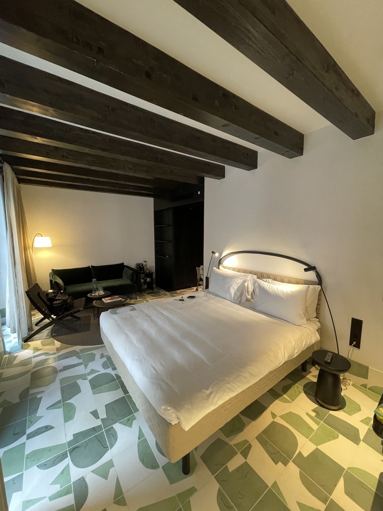 A bed under a dark beamed ceiling on a floor of green and white terrazzo