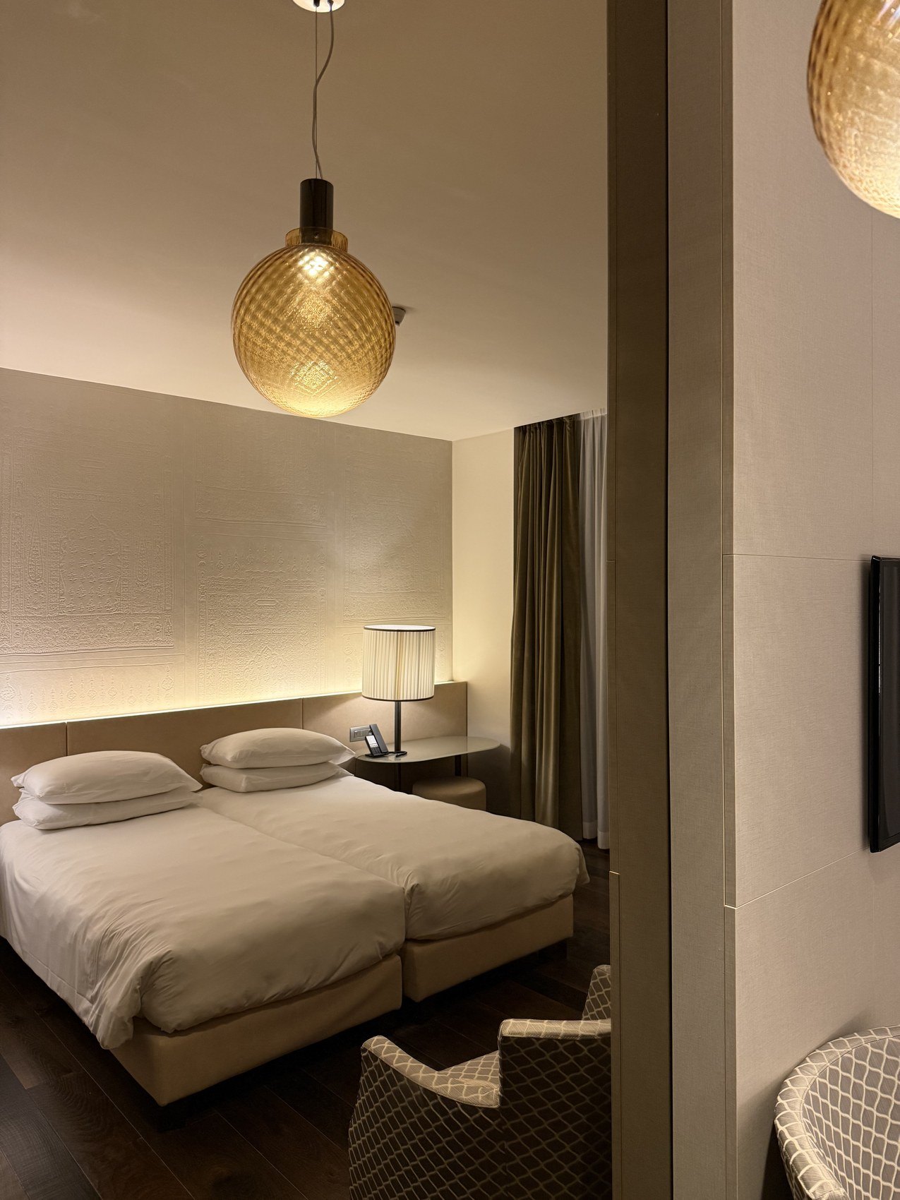 A bed in a pale room under a woven amber globe lamp, with a lit alcove behind it