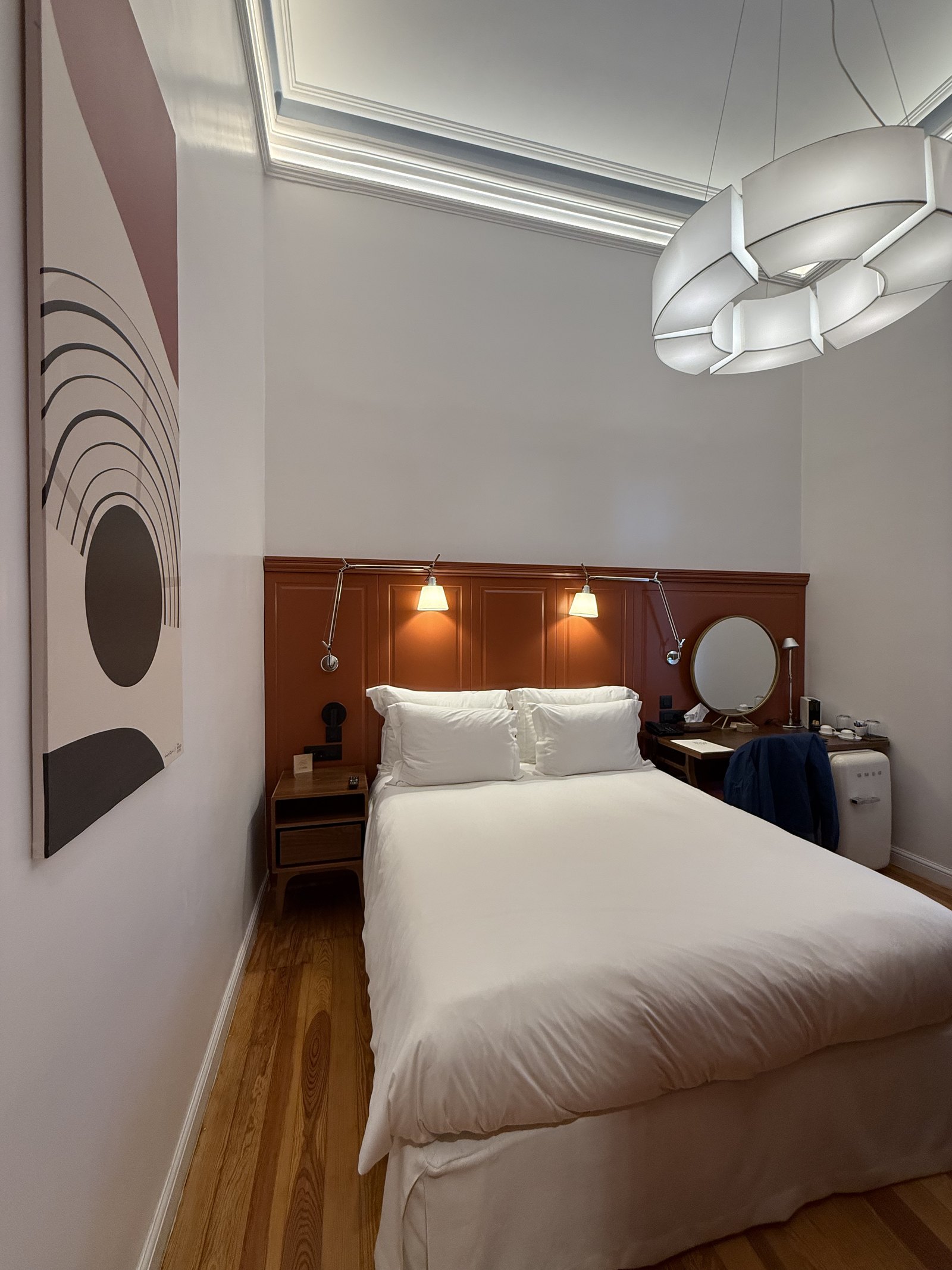 A bed with white linen and a timber headboard under a large print of arcs and circles, with a reading lamp on each side and a round mirror by the desk