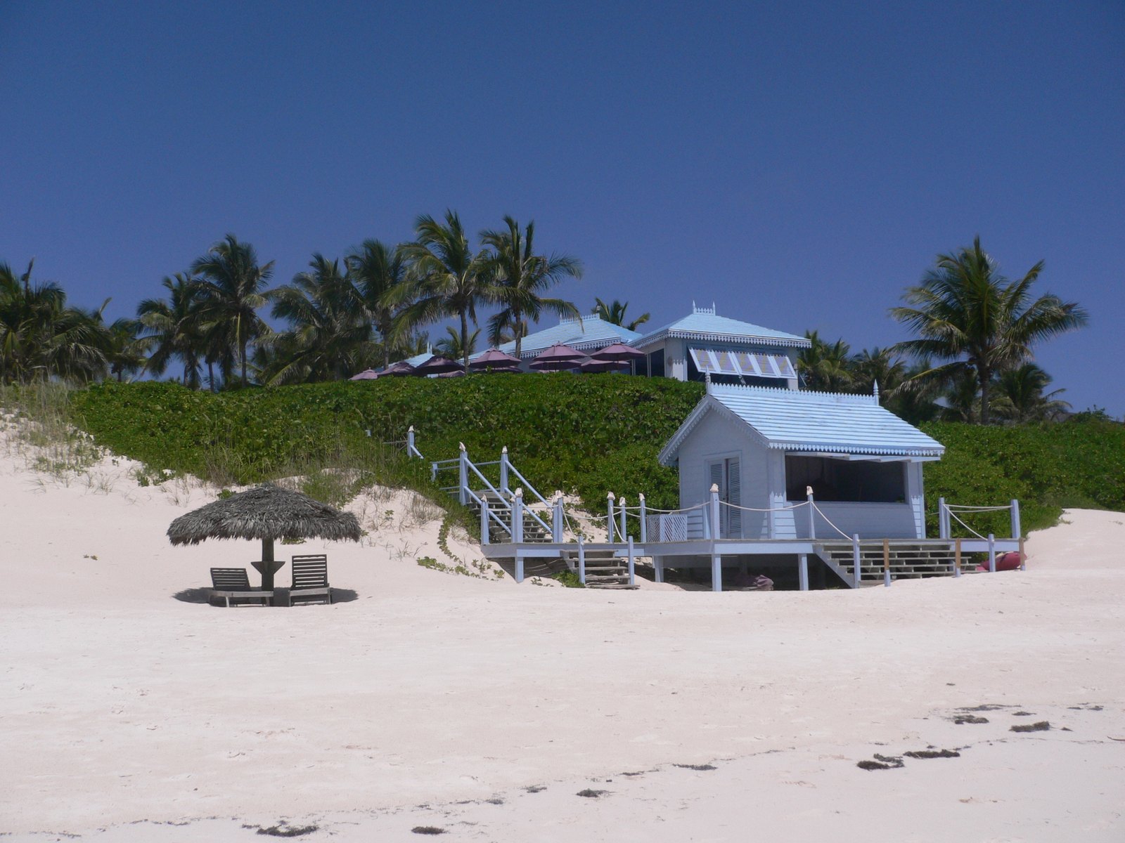 A pale blue beach house with a fretted roof on the dune, a thatch parasol on the sand in front of it