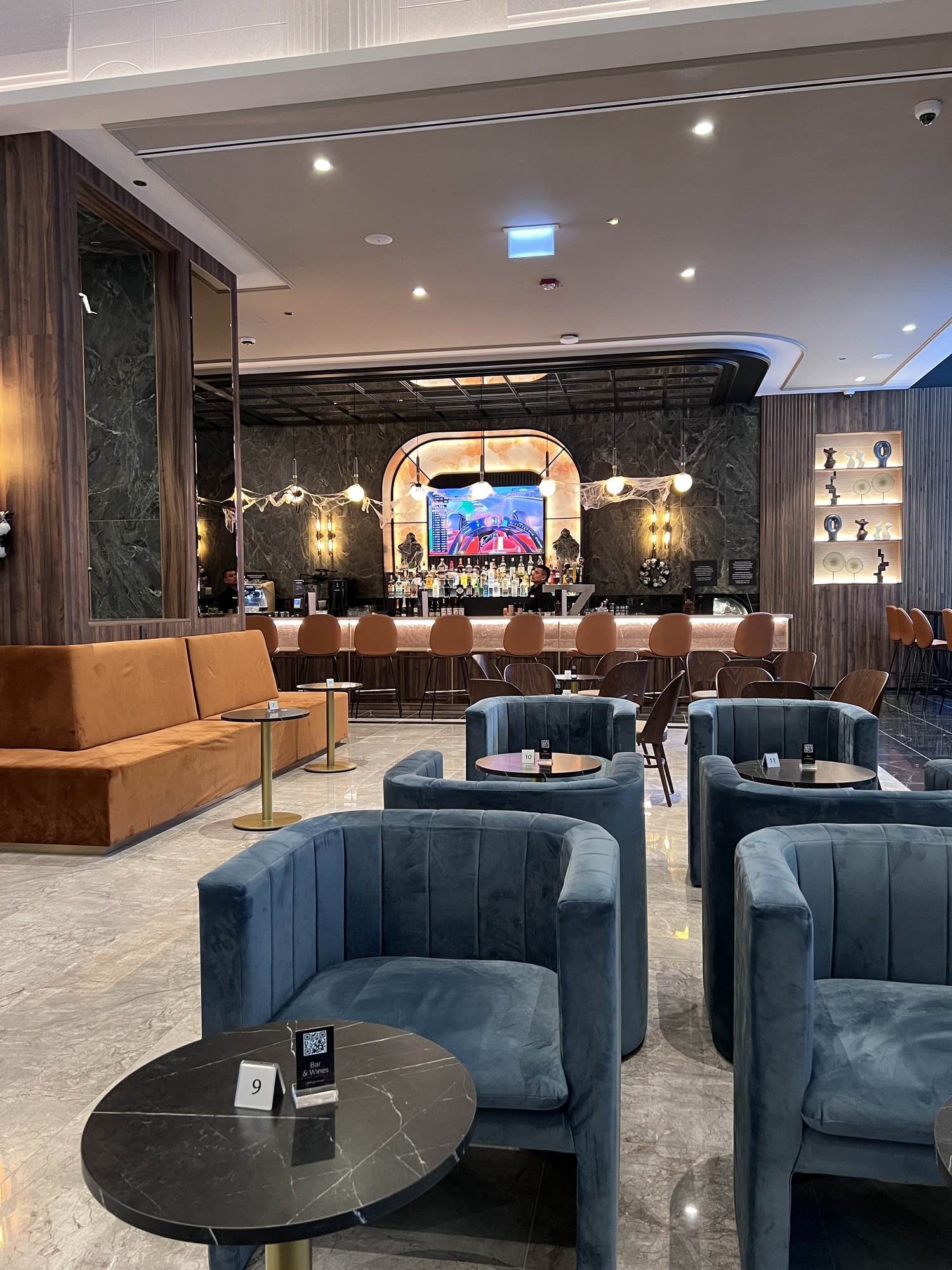 A hotel lounge with blue velvet tub chairs round black marble tables, a long tan banquette along one wall and a lit bar across the far end