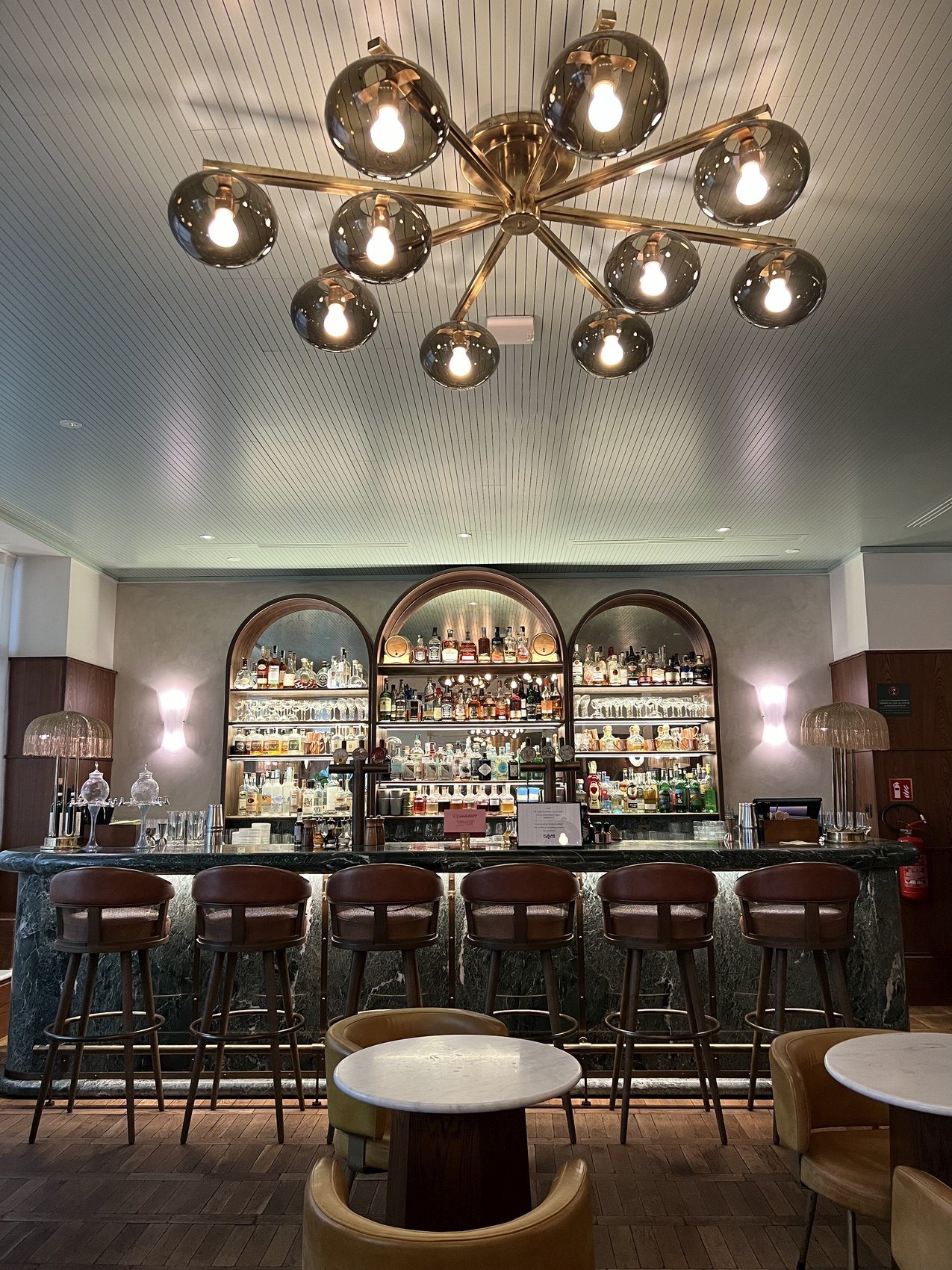A bar lit by a brass chandelier of glass globes, with arched shelves of bottles behind it