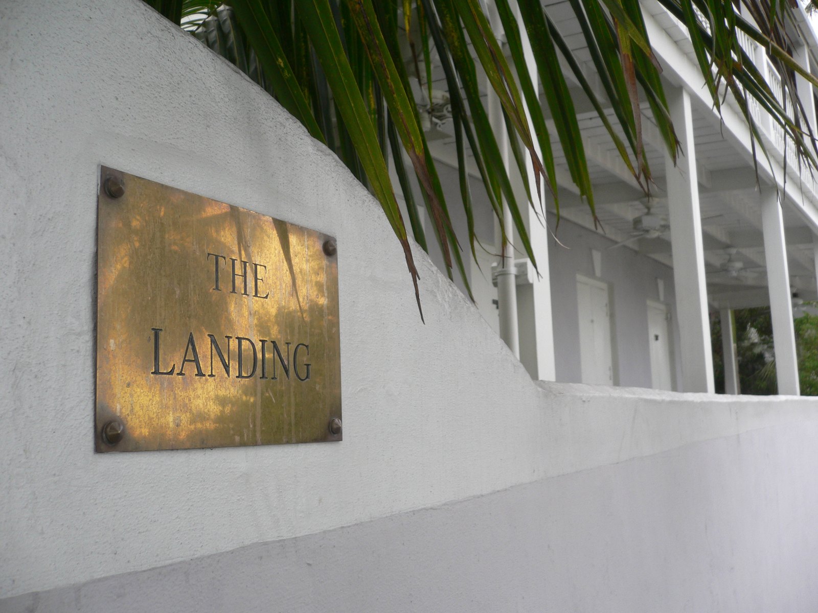 A weathered brass plaque reading The Landing on a white rendered wall, beside a white colonnade under palm fronds