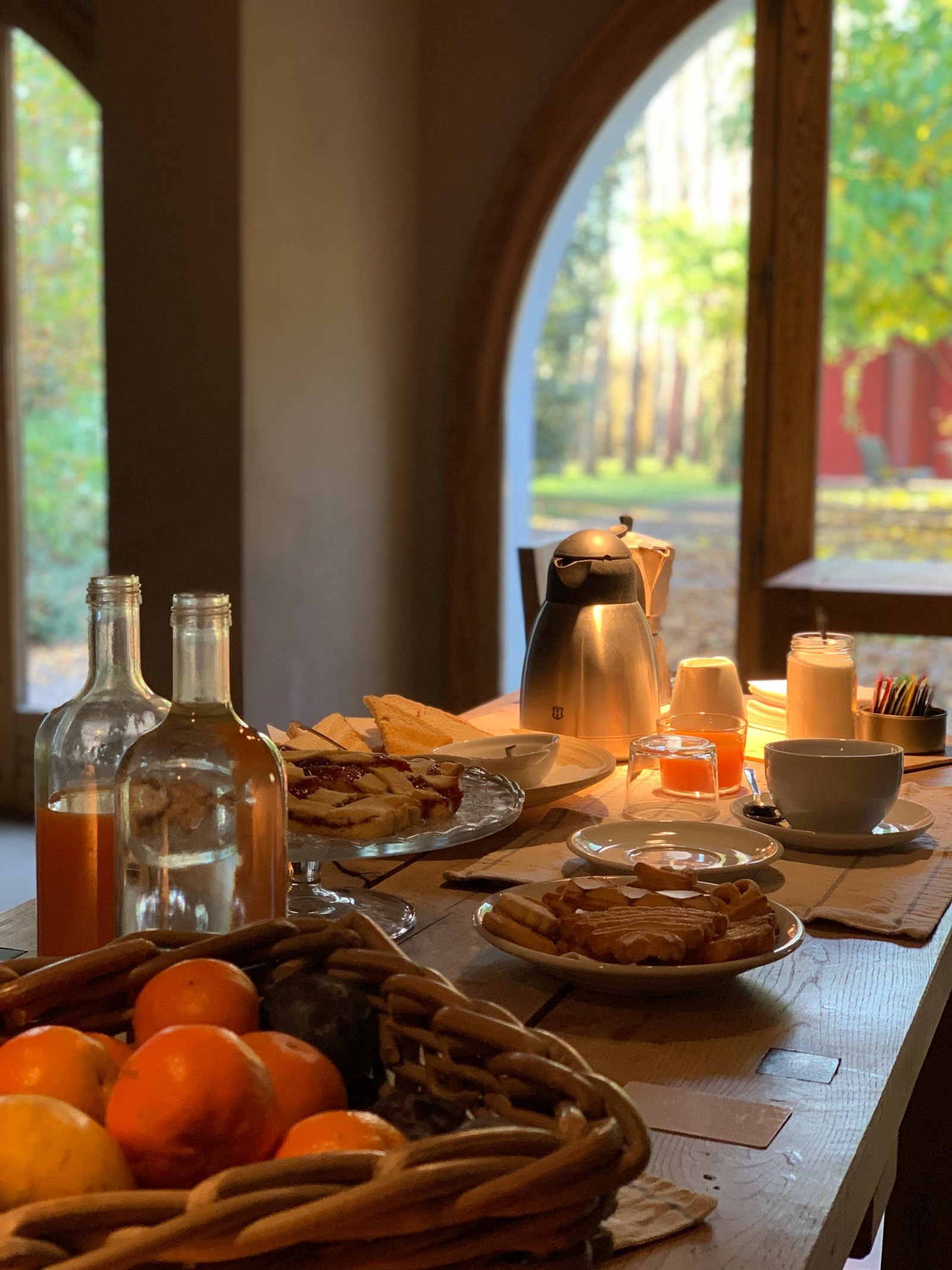 A long timber table laid for breakfast beside a tall arch, with a jam crostata on a glass stand, a plate of biscuits, a steel thermos and a basket of persimmons, autumn woods outside
