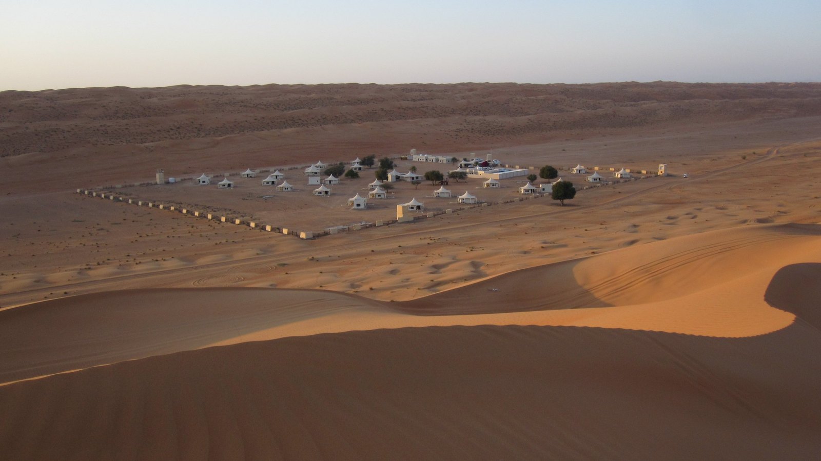 A fenced camp of white tents seen from the top of a dune at dusk, the sand running down towards it