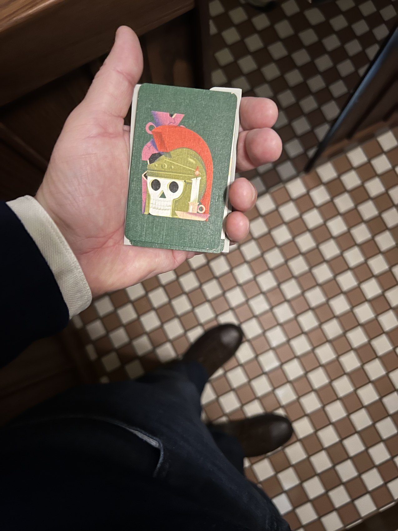 A hand holding a small illustrated card over a brown and white checkerboard floor