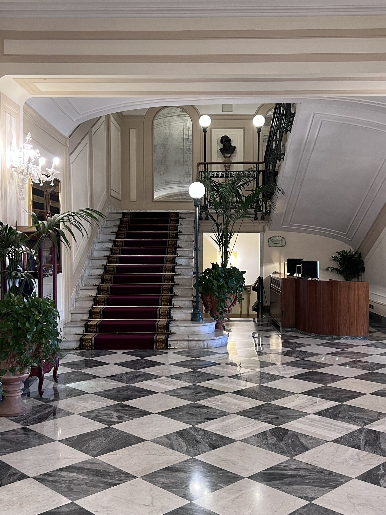 A hall on a black and white chequered marble floor with a red carpeted staircase and globe lamps