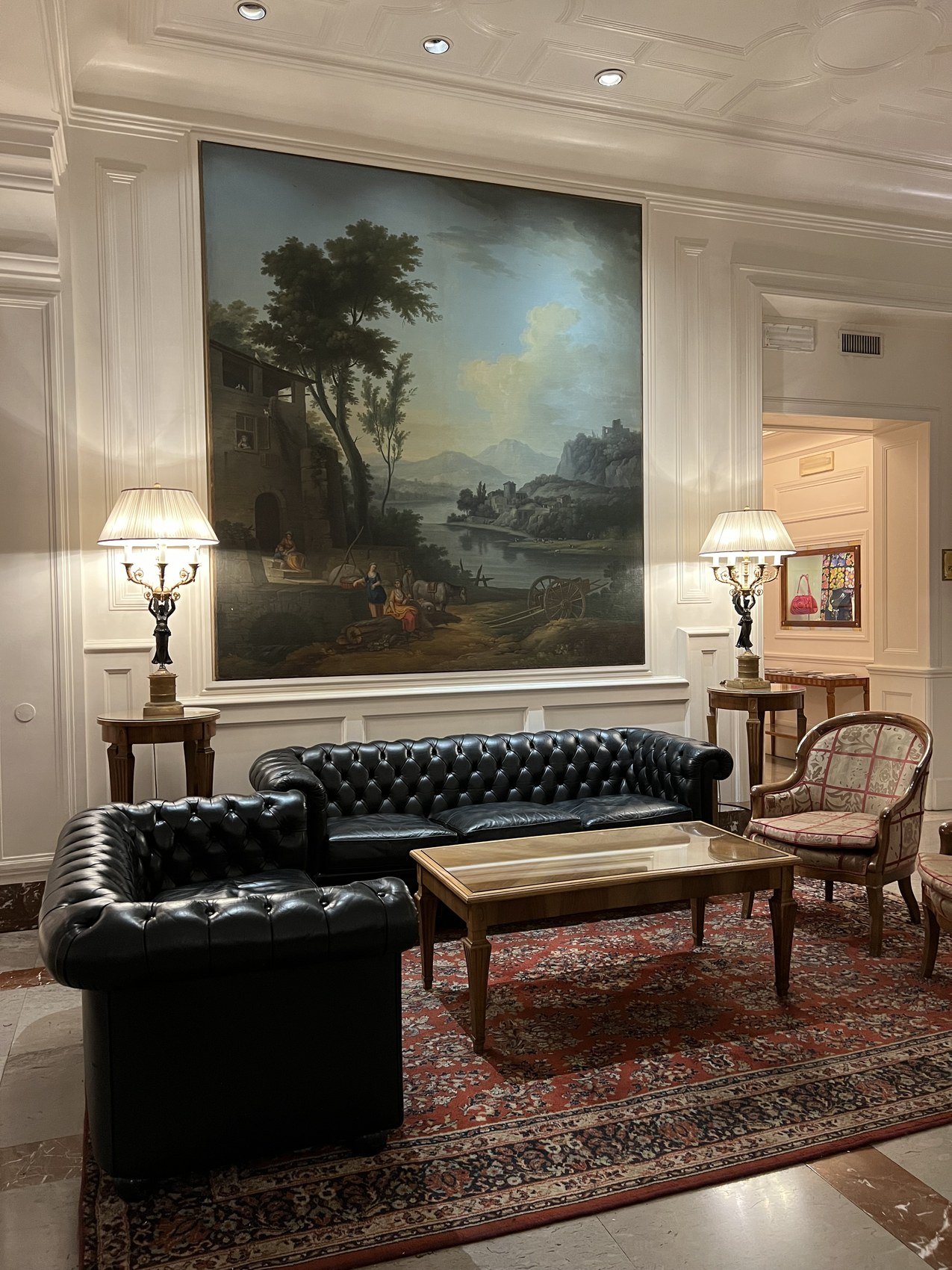A black buttoned sofa below a large landscape painting, on a patterned rug under a moulded ceiling
