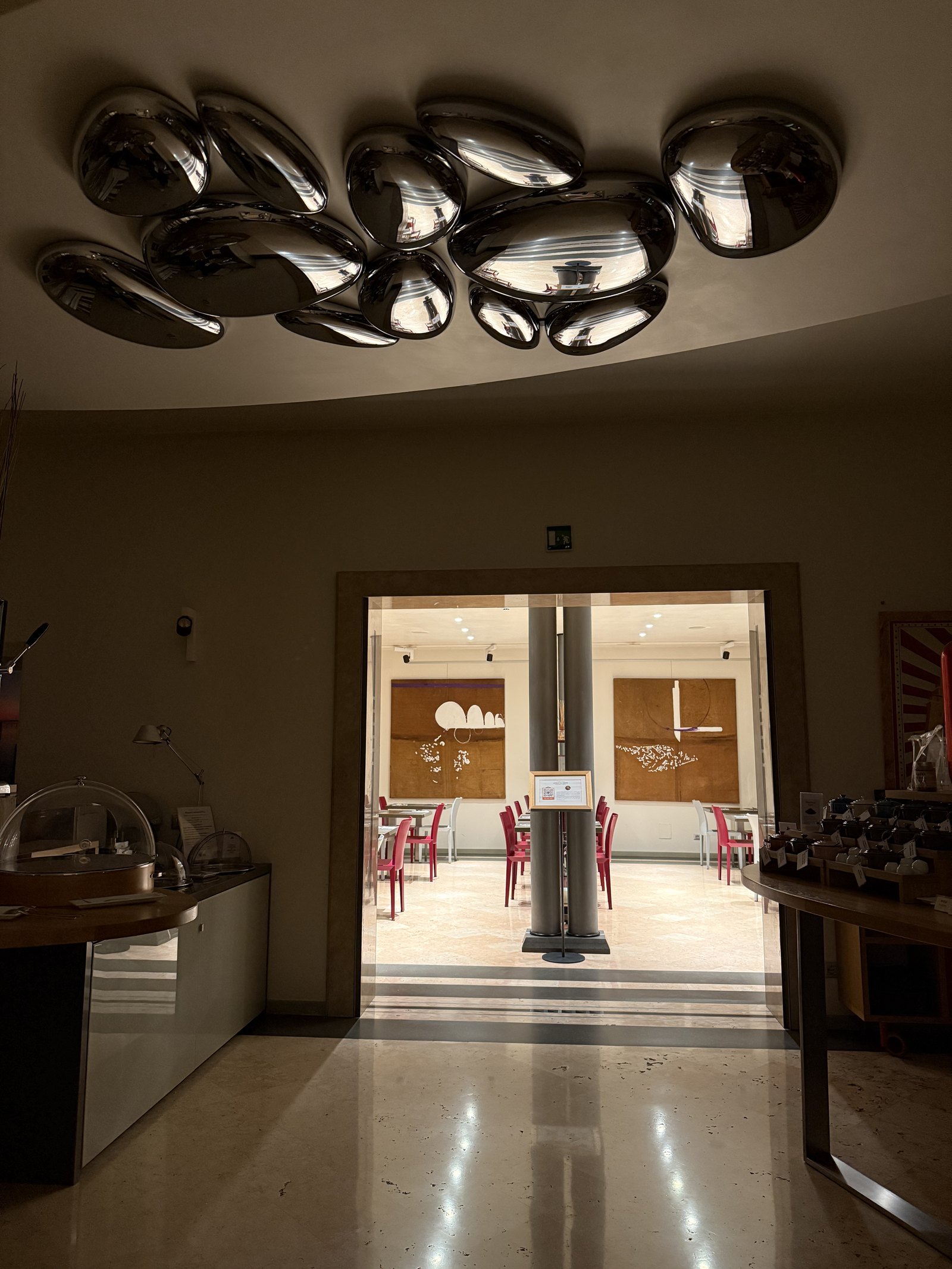 A cluster of polished chrome blobs mounted on the ceiling of a lobby, with a lit doorway through to a room of red chairs beyond