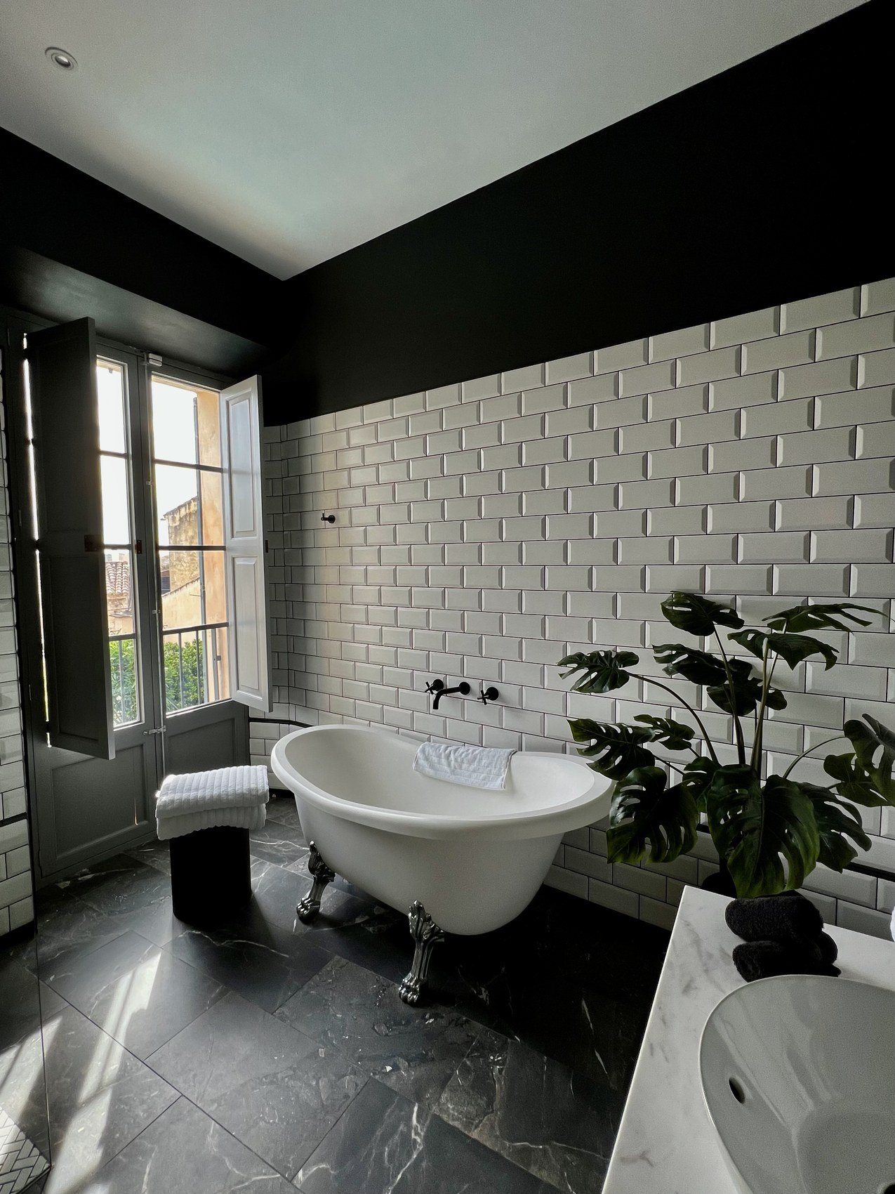 A clawfoot bath against white brick tiling in a dark green room with plants at the window