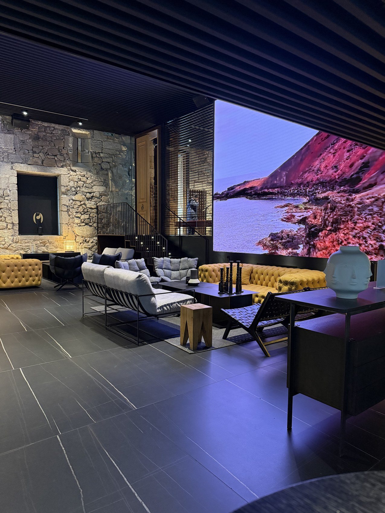 A dark stone lounge with buttoned yellow sofas and a lit screen showing a red cliff and the sea