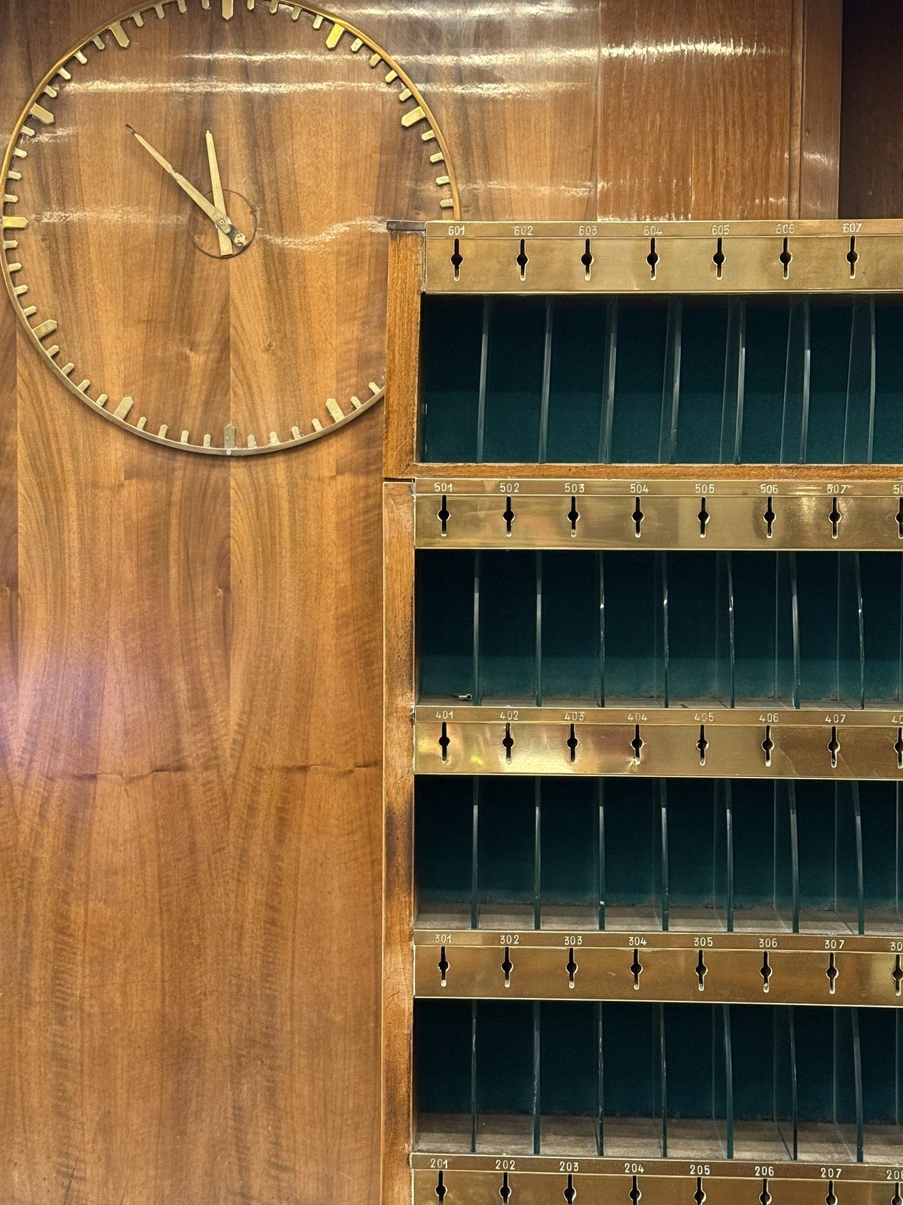 A brass clock face on walnut beside a wall of green and brass key boxes