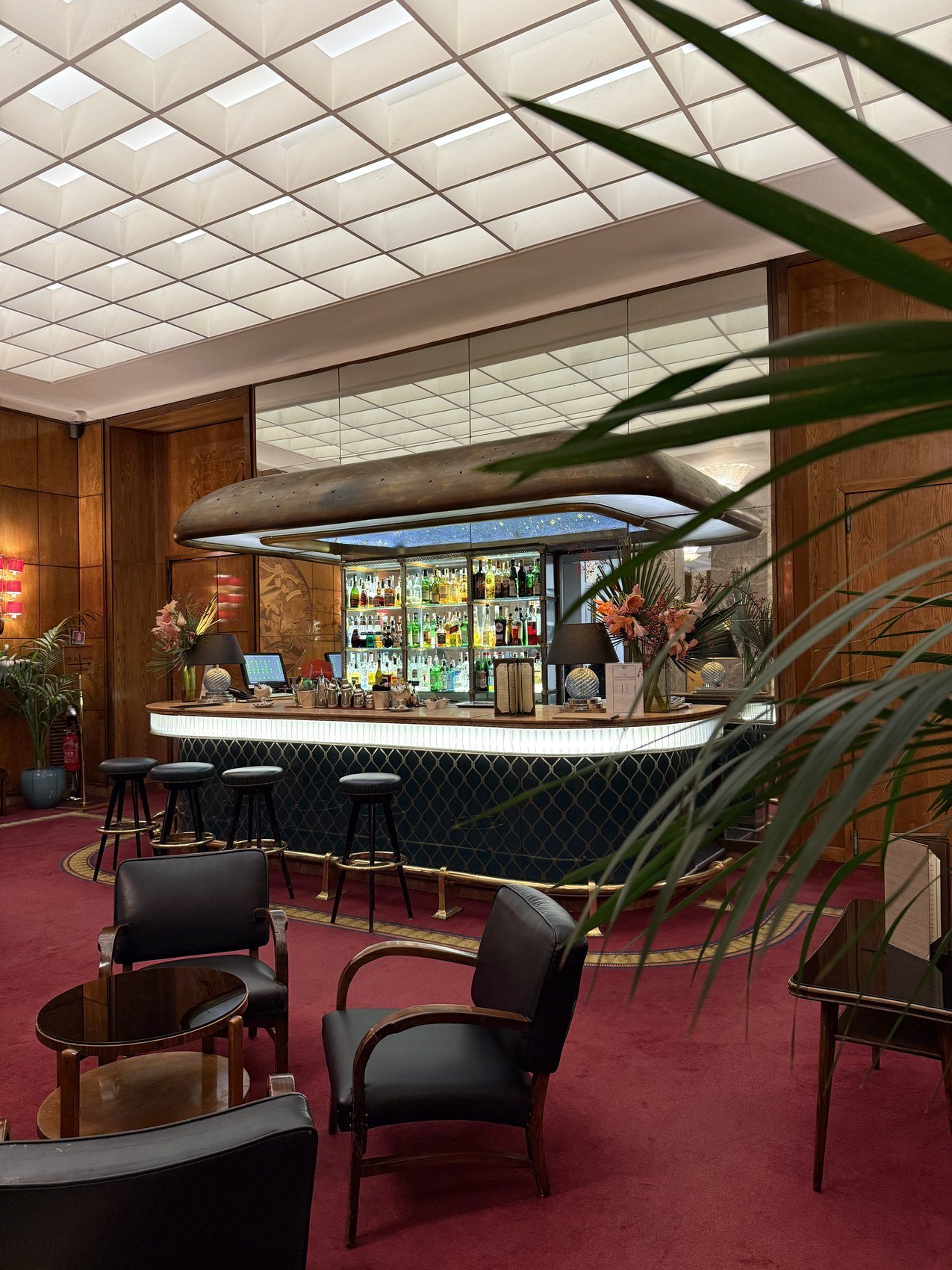 A bar under a white coffered ceiling with a red carpet and dark leather chairs