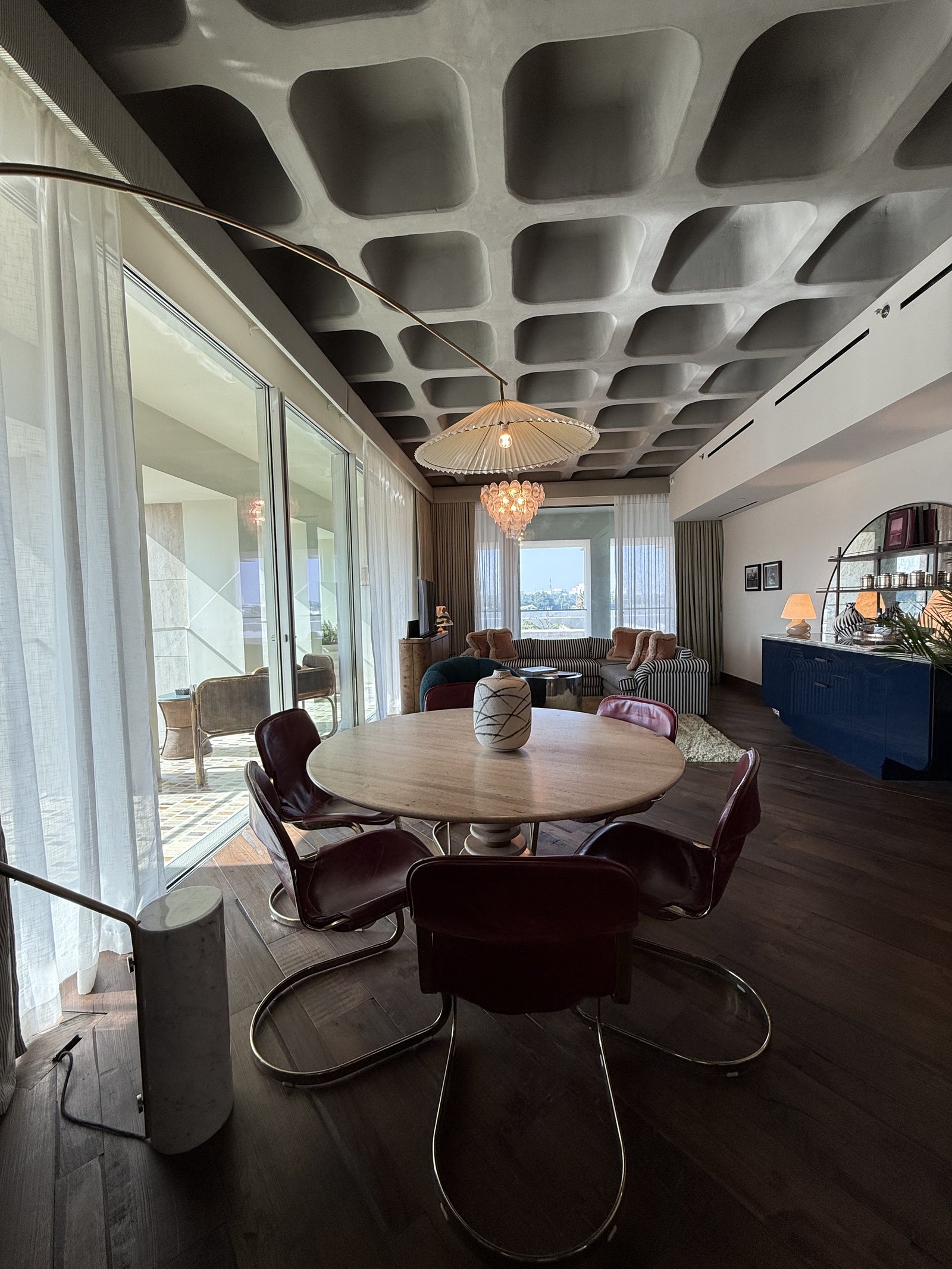 A round timber table and chrome-framed chairs in a suite below a deep coffered concrete ceiling, the city beyond the curtains