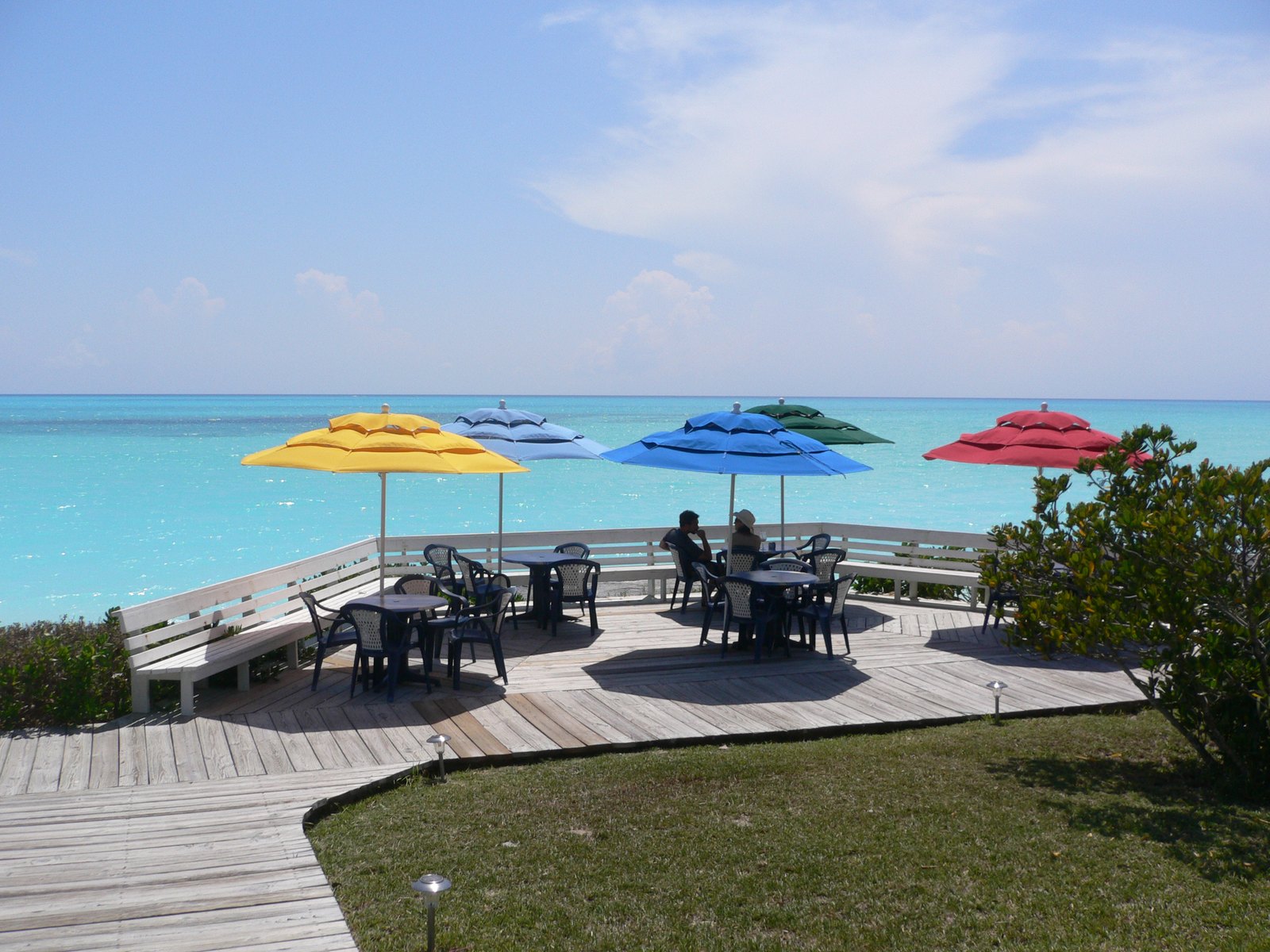 Yellow, white, blue, green and red parasols over tables on a timber deck above the turquoise shallows